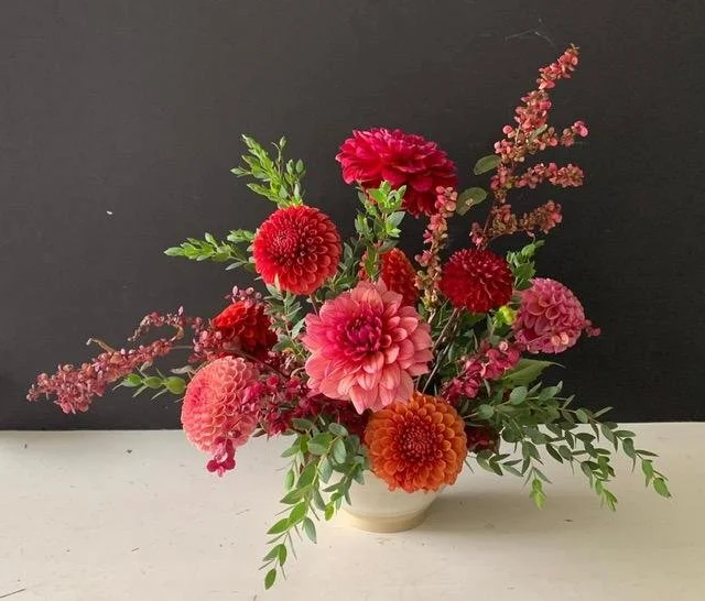 A floral arrangement with red and pink dahlias, fuchsia, and pink blossoms in a white vase against a black background.