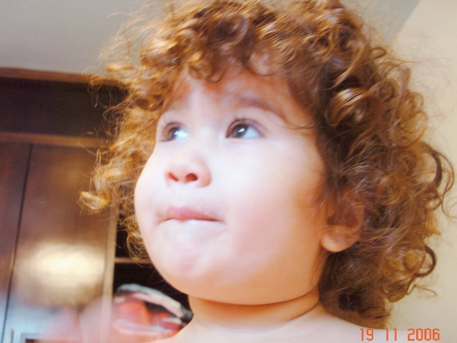 A young child with curly red hair looking slightly upward, with soft lighting and a blurred background.