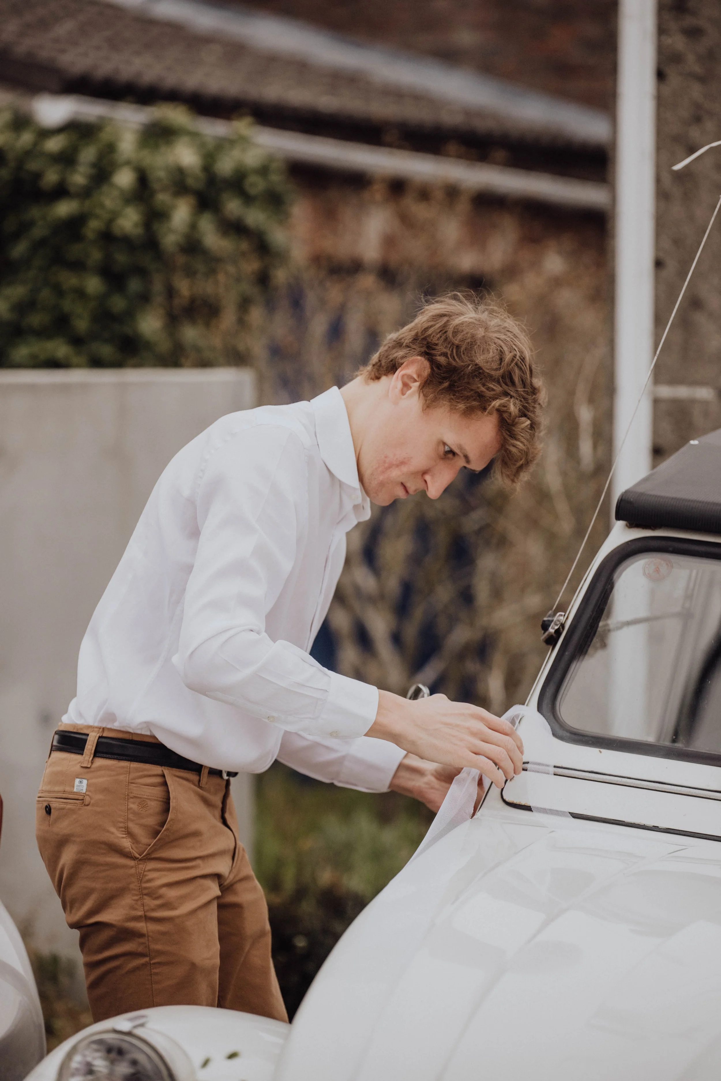 Un jeune homme aux cheveux bouclés, vêtu d'une chemise blanche et d'un pantalon marron, est en train de préparer une 2CV blanche pour un mariage.