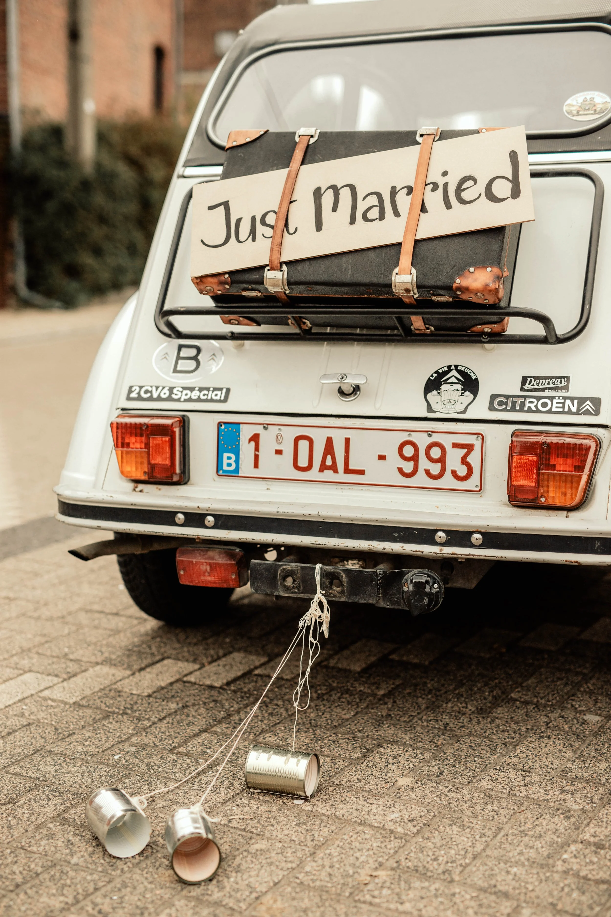 L'arrière d'une Citroën 2CV blanche d'époque, décoré d'autocollants et d'une pancarte sur laquelle on peut lire « Just Married », fixée à une vieille valise à l'arrière. Trois boîtes de conserve vides sont attachées au pare-chocs de la voiture.