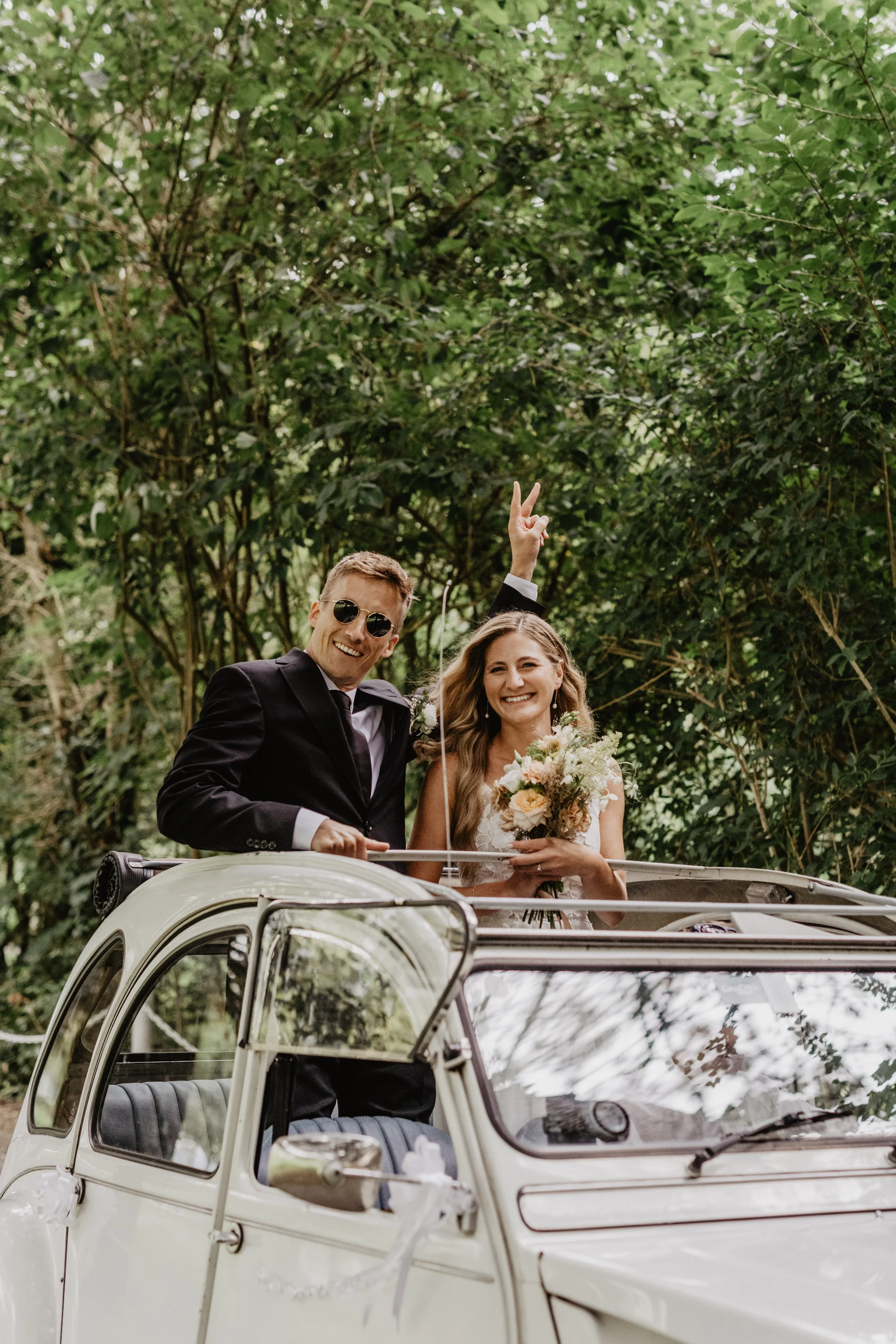 Un couple souriant, en tenue de mariage, profite d'une balade en 2CV blanche d'époque en plein air, au milieu de la verdure. Le marié porte des lunettes de soleil noires et un costume noir, tandis que la mariée tient un bouquet de fleurs et sourit.
