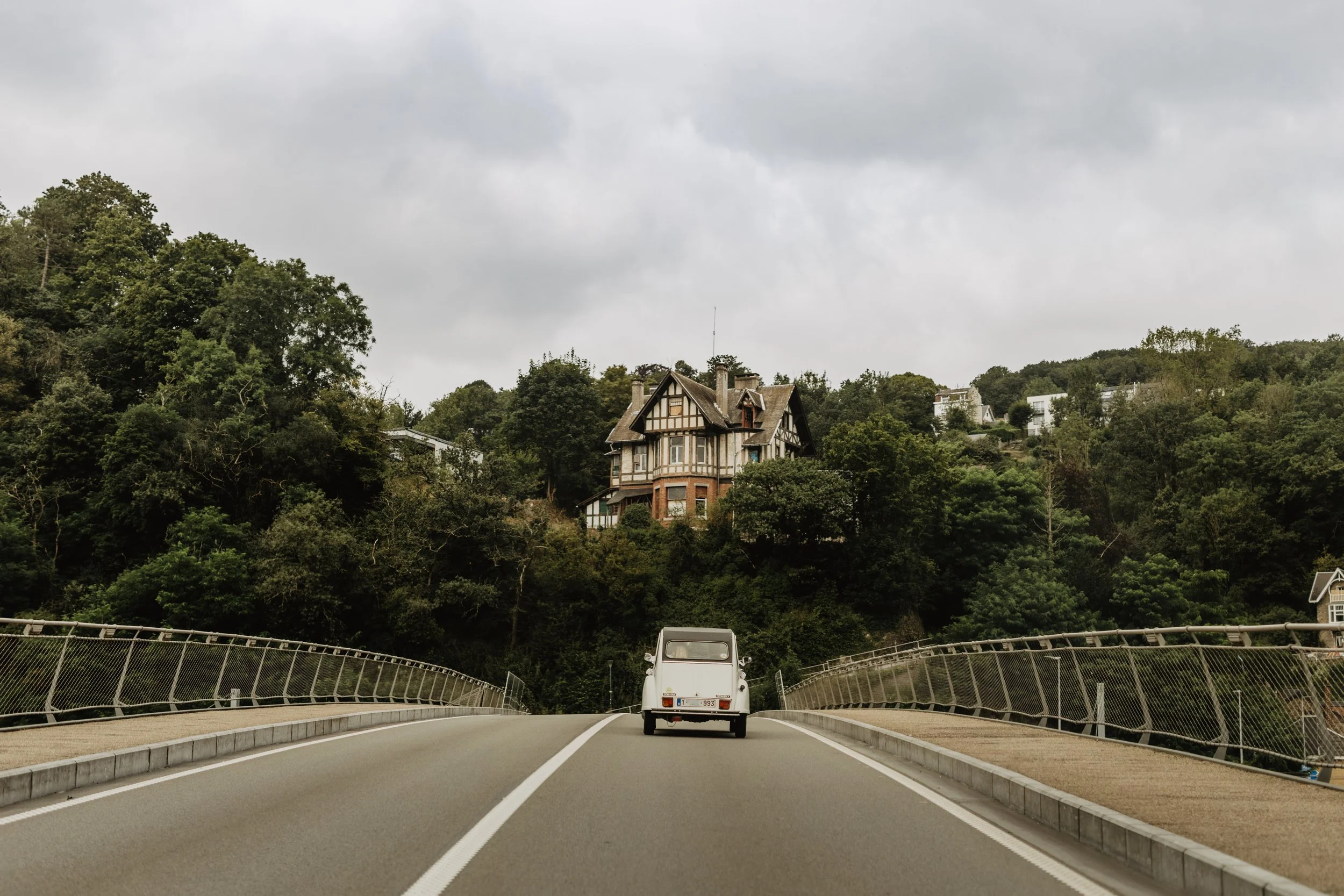 Une voiture ancienne roulant sur une route de montagne sinueuse, entourée d'arbres verdoyants et de maisons à flanc de colline, sous un ciel nuageux.