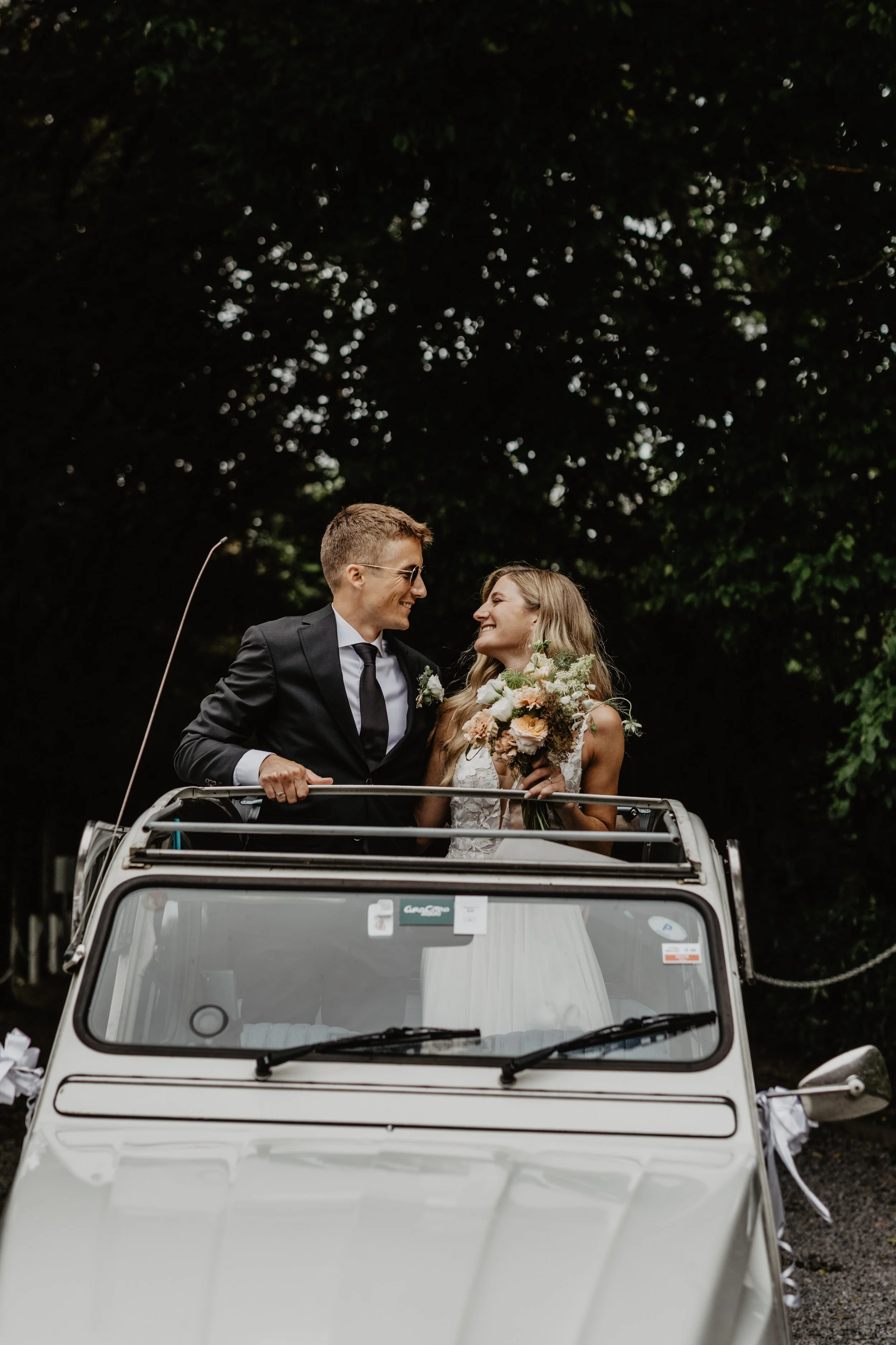 Un couple de jeunes mariés qui s'échangent un sourire à bord d'une voiture blanche d'époque, un bouquet de fleurs à la main, dans un décor en plein air avec des arbres verdoyants en arrière-plan.