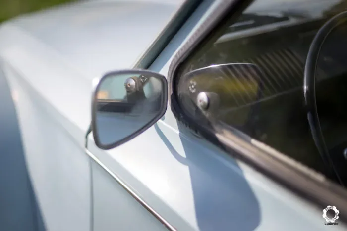 Gros plan sur le rétroviseur latéral d'une Dyane bleue de collection, montrant la vitre du véhicule et le reflet du ciel.