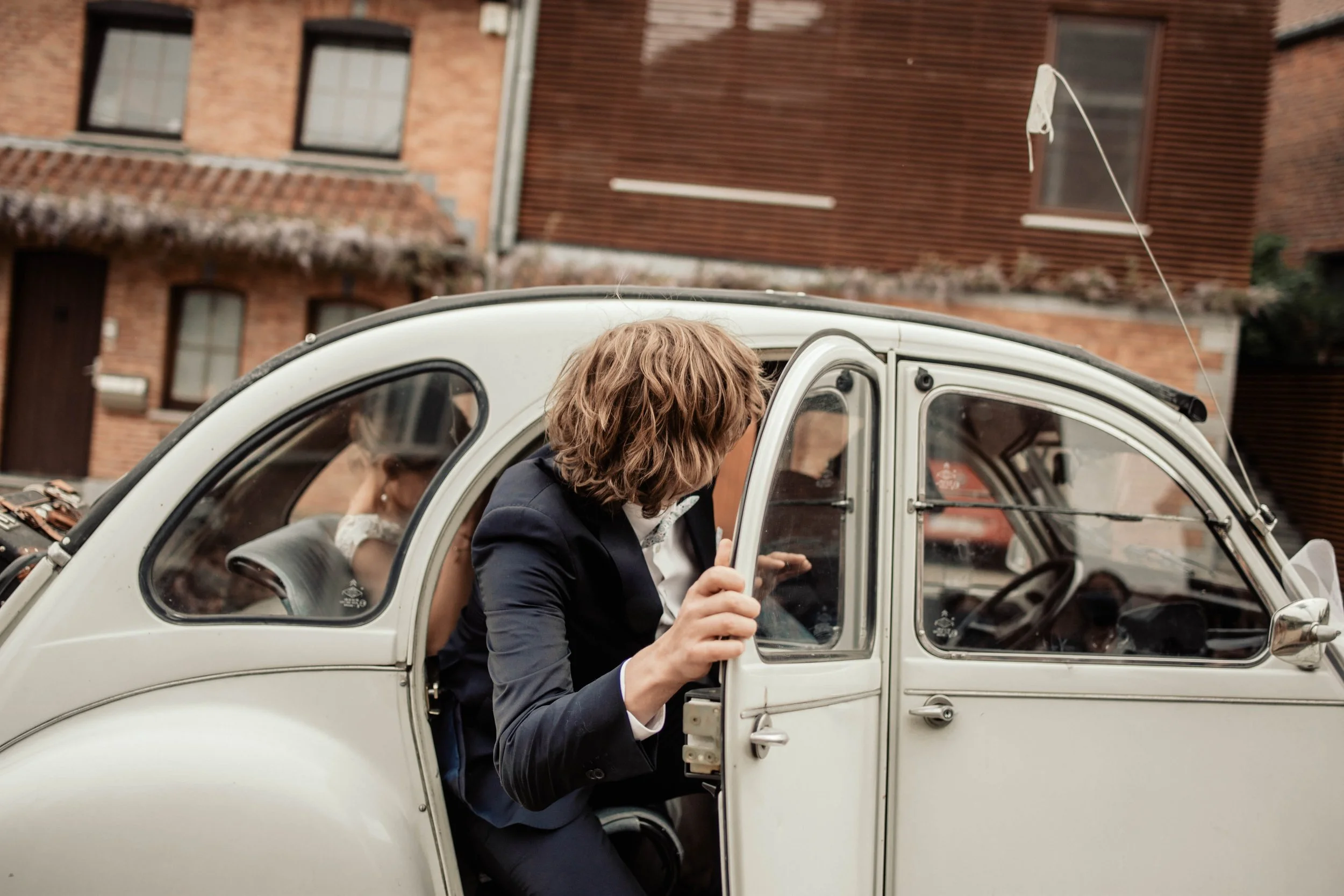 Une personne aux cheveux courts et bouclés, vêtue d'un costume noir, monte dans une voiture blanche d'époque.
