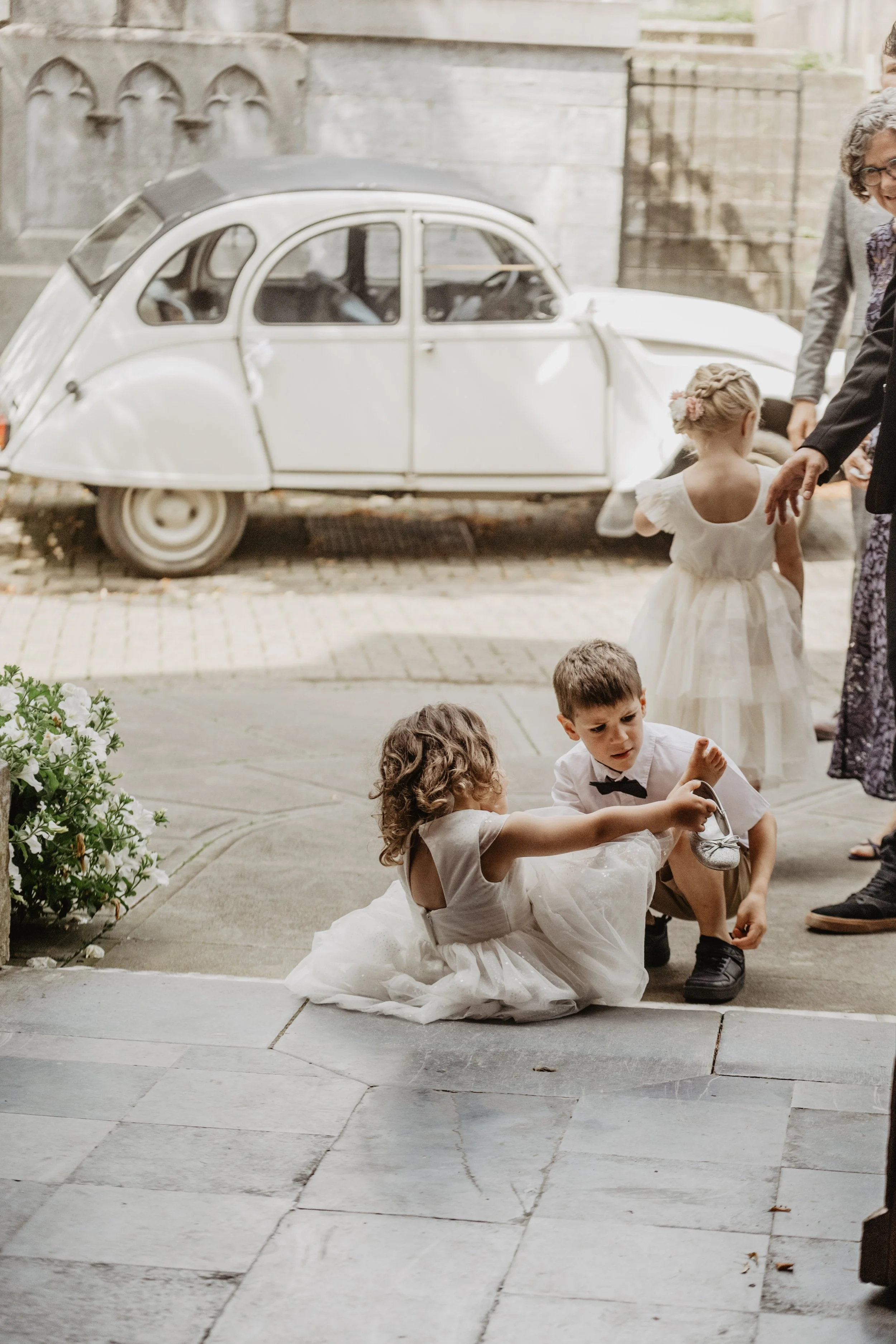 Deux enfants en tenue de cérémonie, une fillette en robe blanche et un garçon en chemise blanche avec un nœud papillon, assis par terre et jouant avec un petit objet, avec une voiture blanche d'époque en arrière-plan, devant un bâtiment en pierre.
