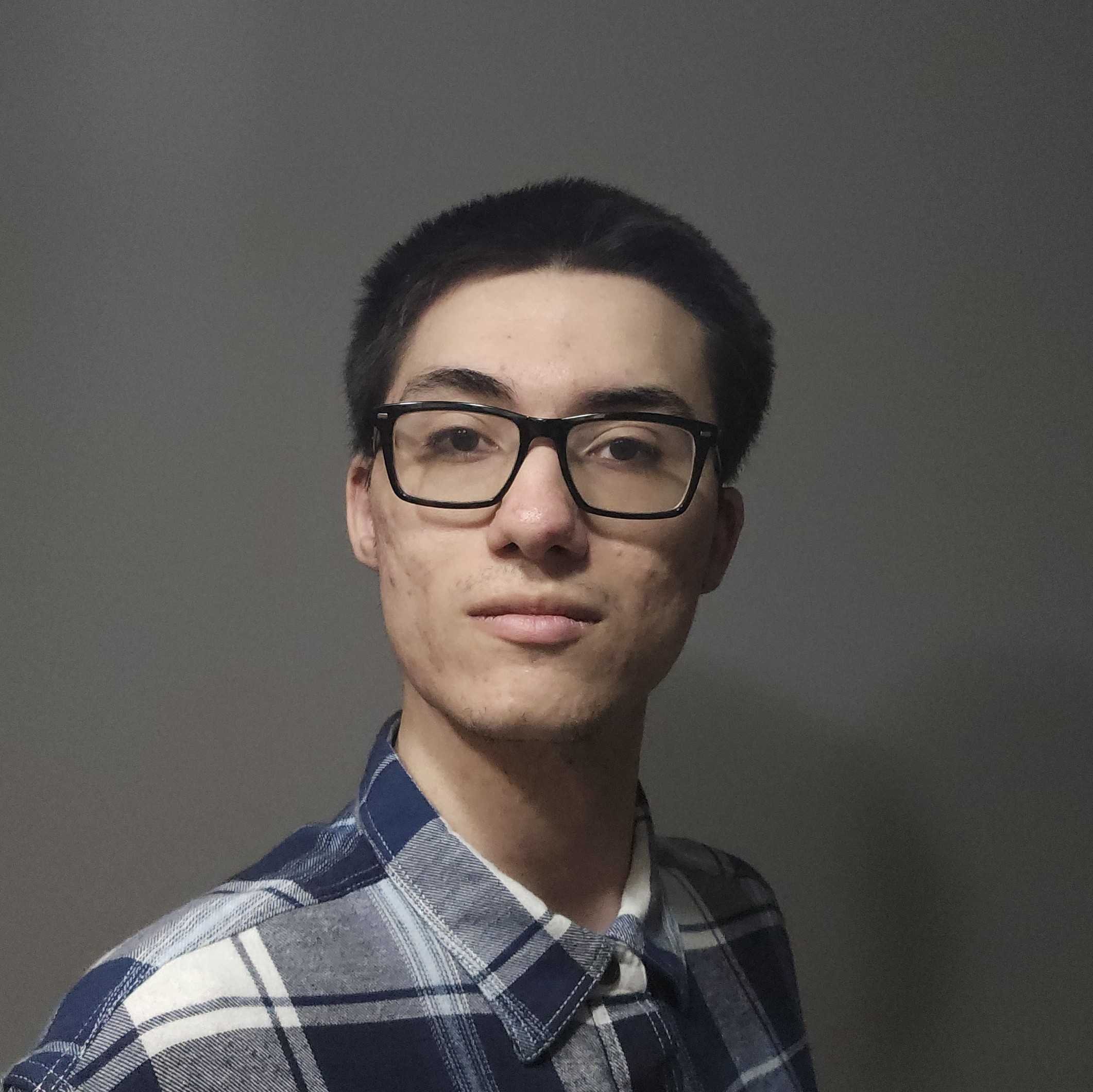 A young man with dark hair, wearing glasses and a plaid shirt, standing in front of a plain gray wall.