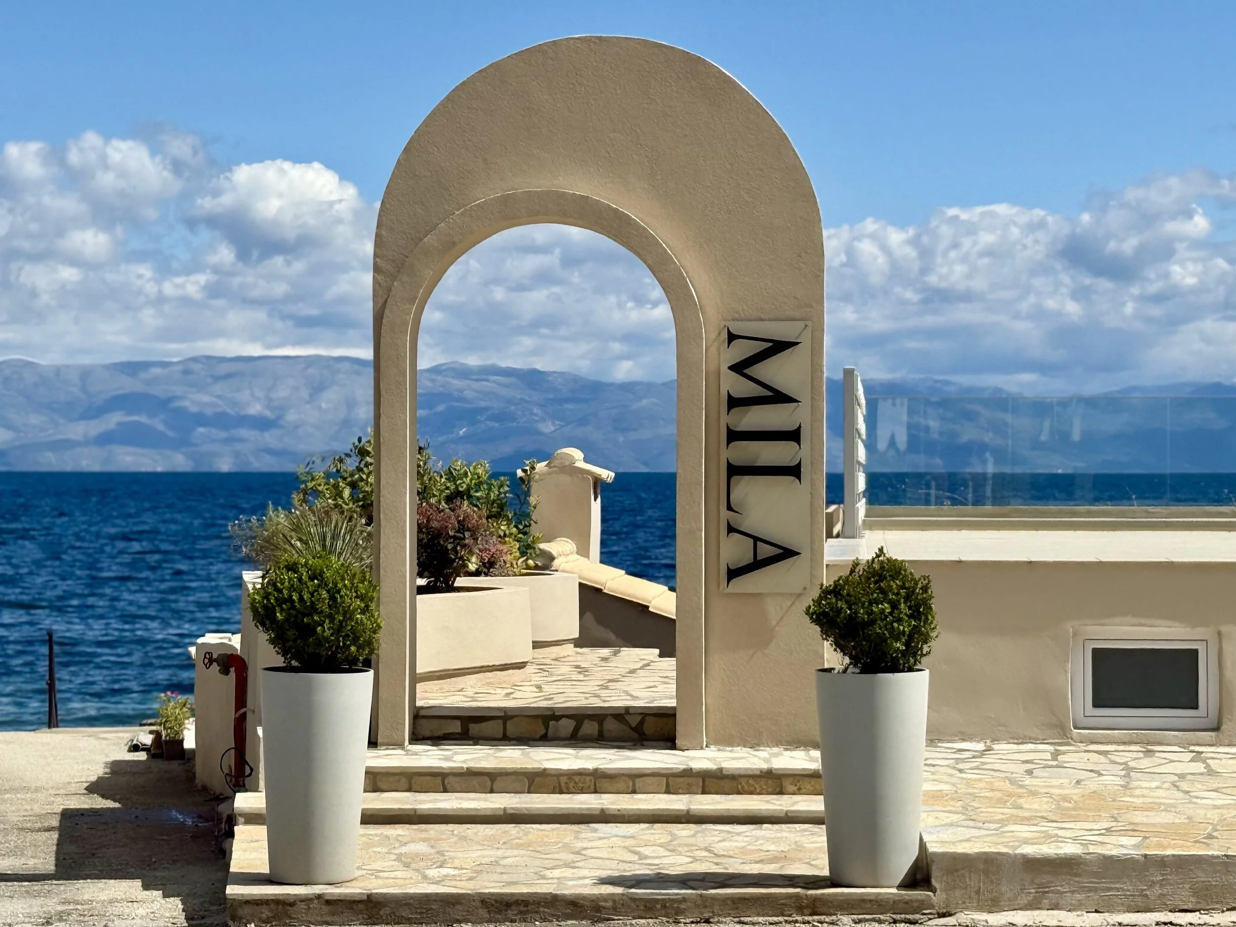 View of the entrance with an archway labeled 'MILA' overlooking the sea with mountains in the distance, blue sky, and scattered clouds.