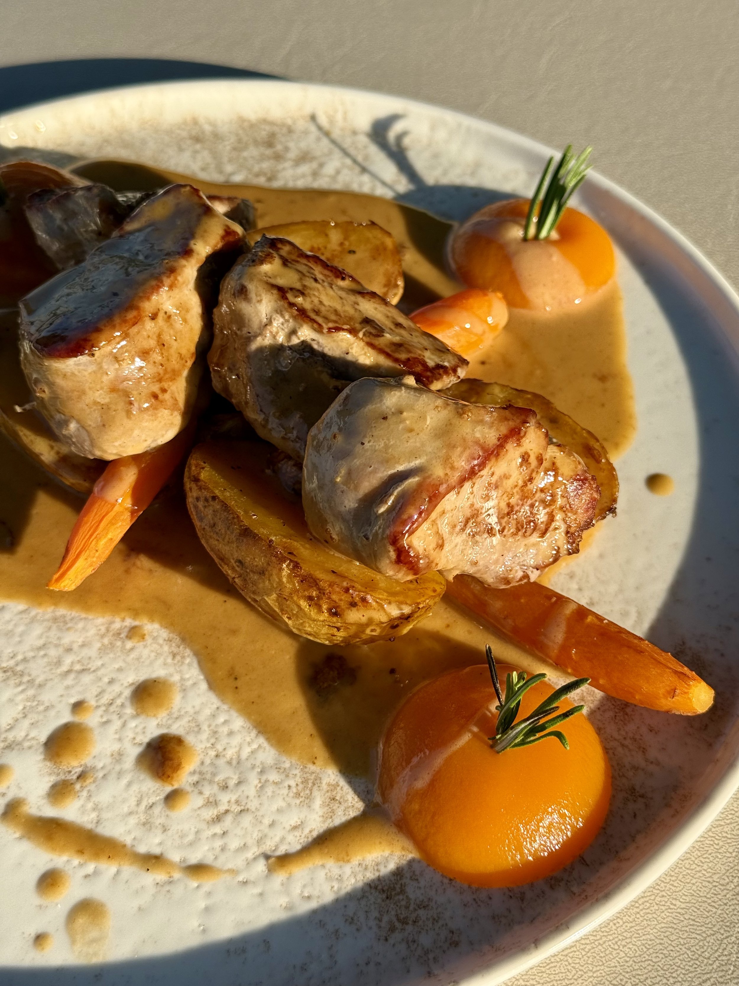 Plate of cooked meat, carrots, potato, and sauce, garnished with green herbs, with drizzles and drops of sauce on the plate.