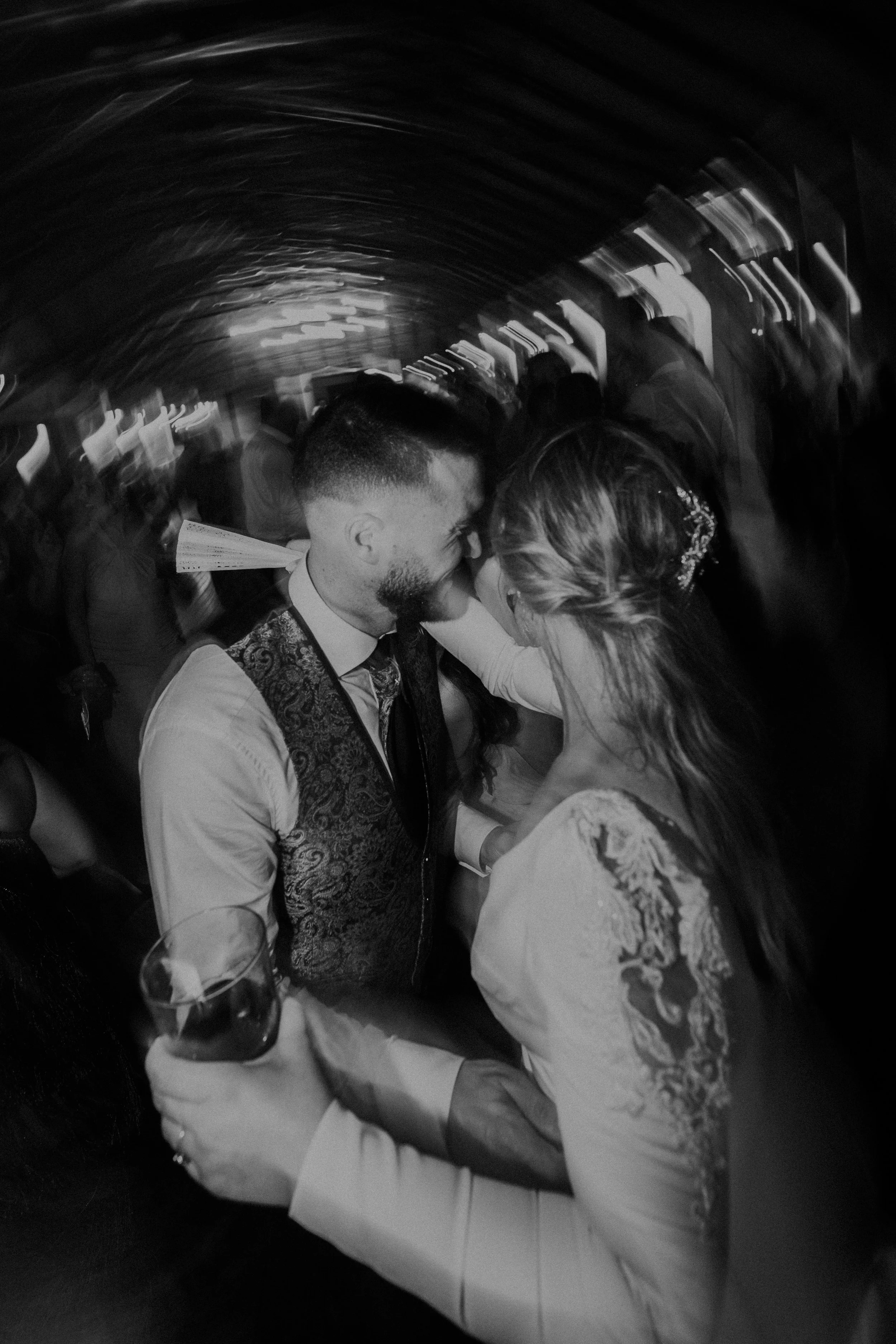 Pareja bailando en una fiesta, ella sostiene un vaso con bebida, ambos en un abrazo cercano en un ambiente oscuro y con movimiento.