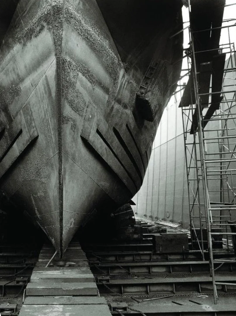Black and white photograph of a vessel's bow in dry dock, surrounded by scaffolding and shipyard structures.