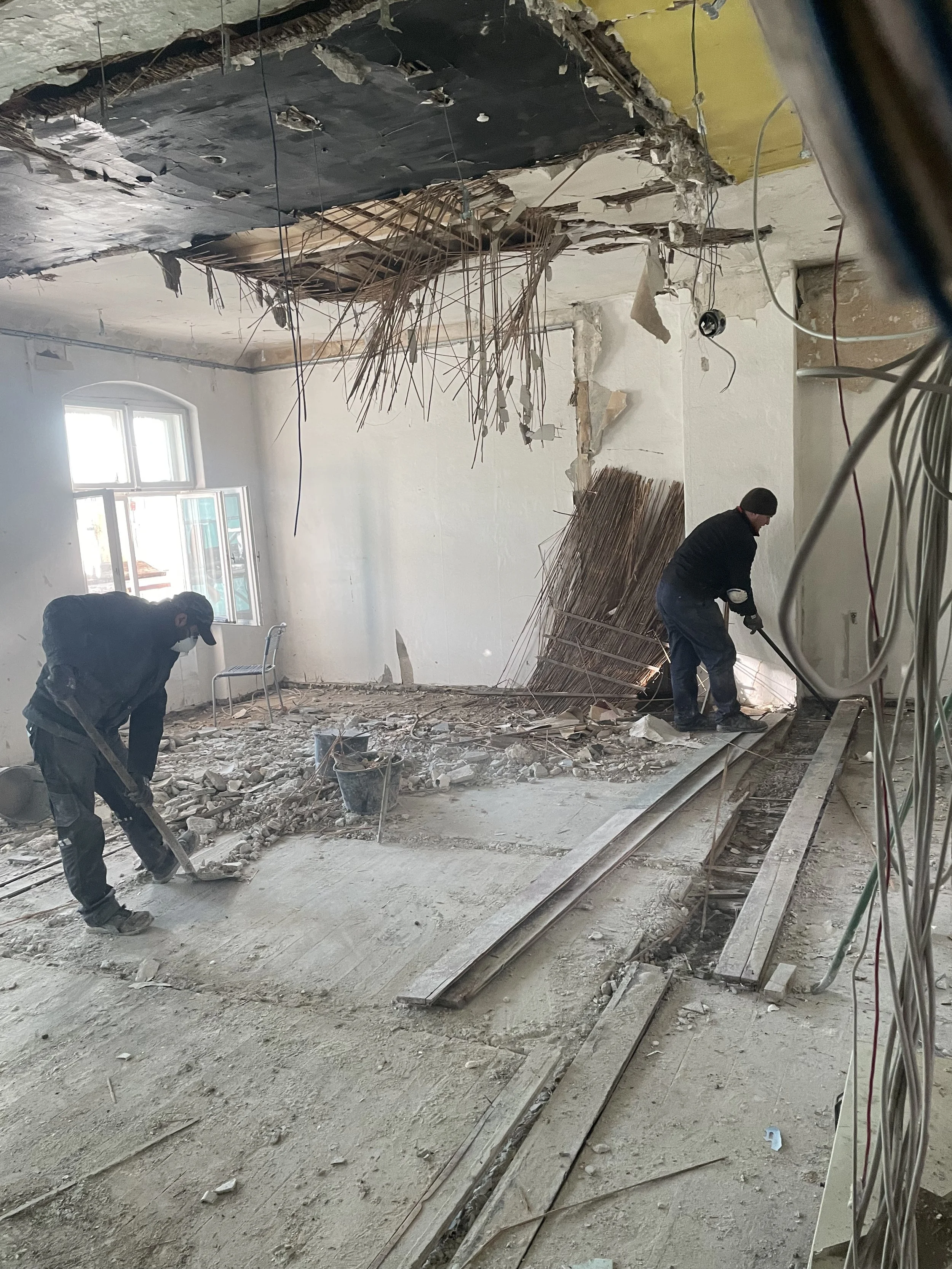Innenraumsanierung mit Abrissarbeiten an Decke und Boden, zwei Arbeiter entfernen Materialien, Trage gerät und Kabel sichtbar, Fenster an der Wand, Staub und Trümmer auf dem Boden.