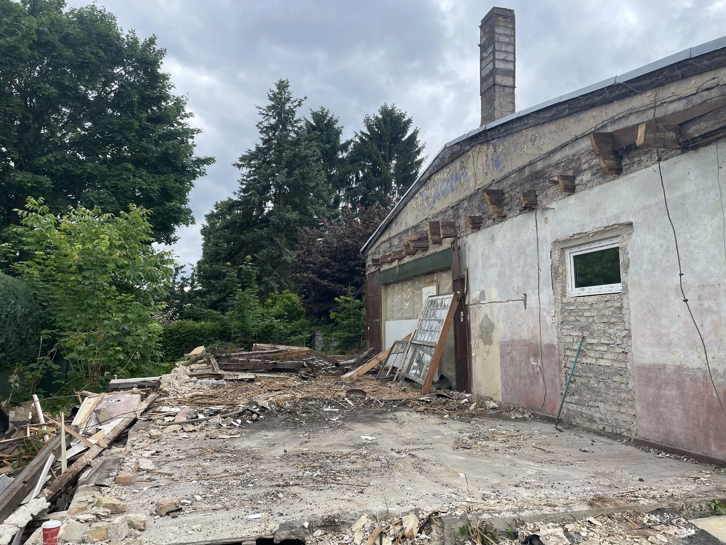 Ein abgebranntes Haus mit zerstörtem Dach und verstreutem Schutt im Vordergrund, umgeben von Bäumen, bei bewölktem Himmel.