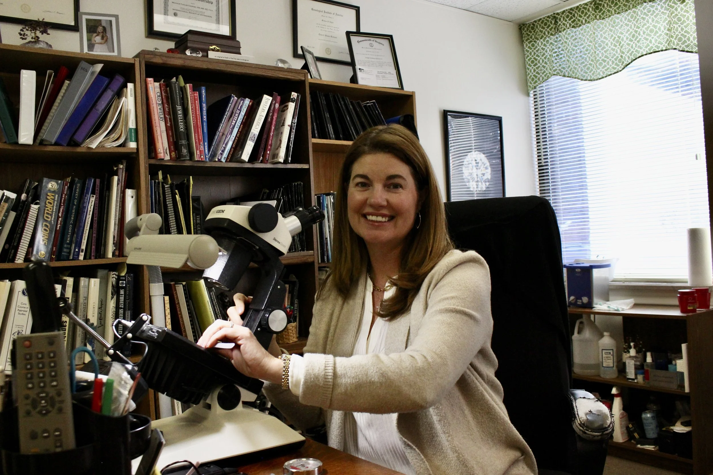 A woman with long brown hair, wearing a cream-colored cardigan, smiling while sitting at a desk with a microscope and scientific equipment in a cluttered office or laboratory with shelves, framed certificates, and a window with blinds