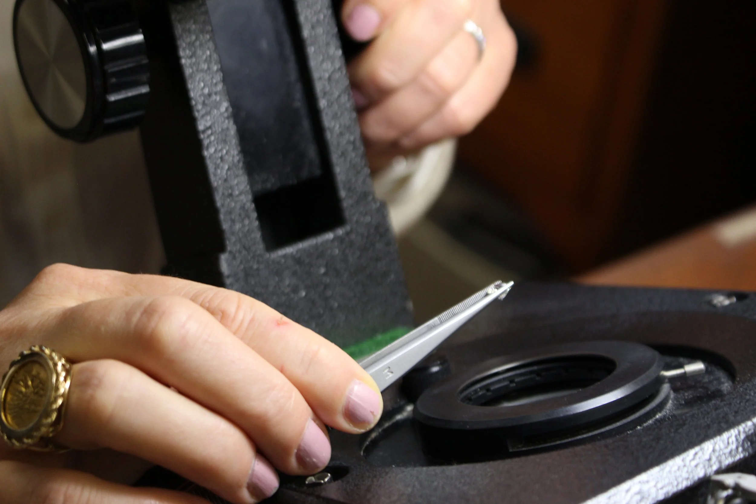 Close-up of a person's hand with gold rings, holding a small metallic screwdriver near a mechanical device, possibly performing repairs.