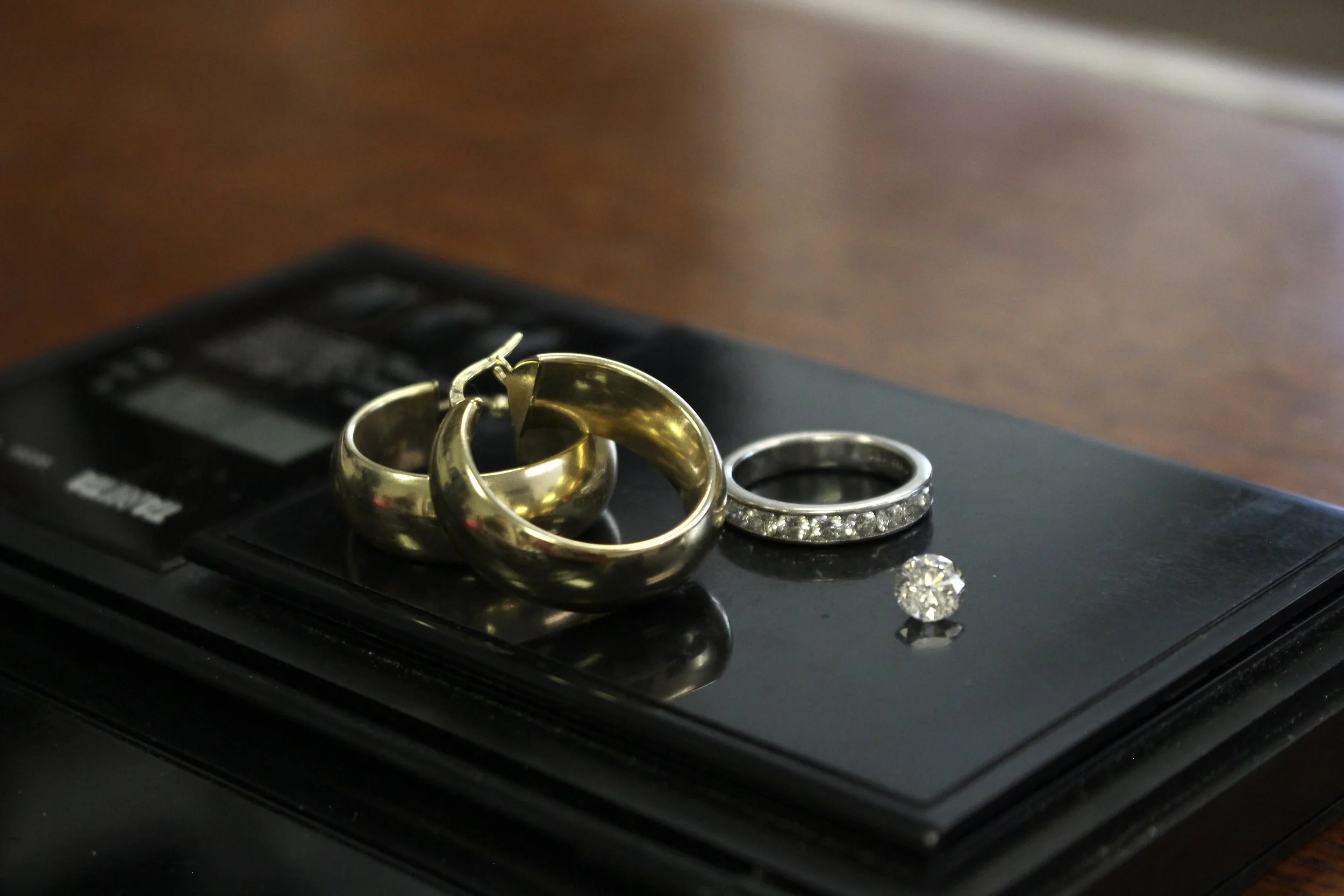 Close-up of gold and silver rings, including a gold wedding band, a diamond engagement ring, and a diamond eternity band, placed on a black electronics device on a wooden surface.