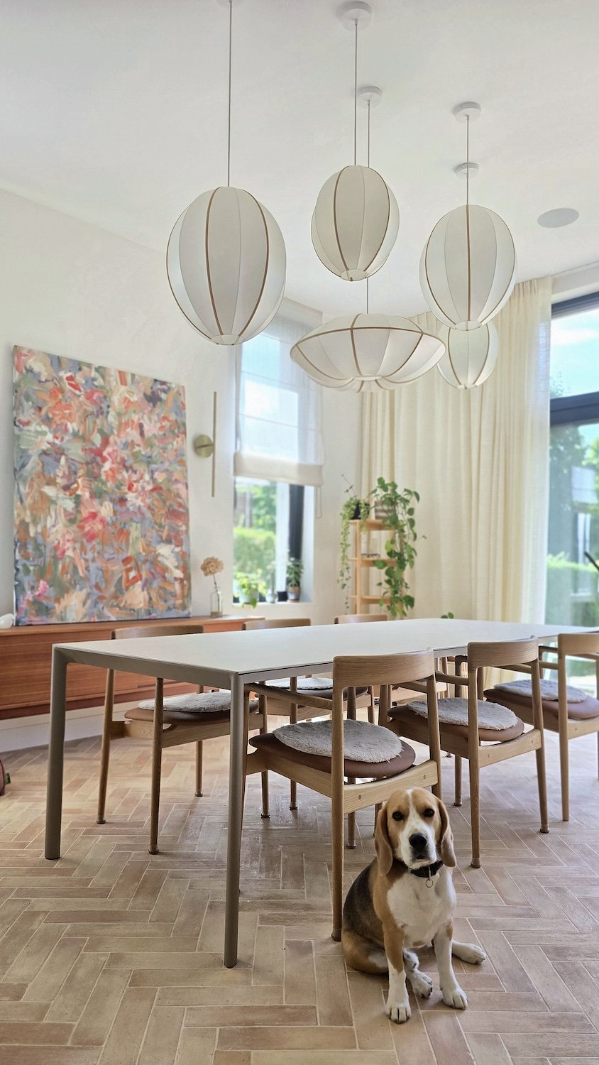Une salle à manger lumineuse avec une grande table blanche, des chaises en bois avec des coussins, des lampes suspendues en forme d'ovale, une grande fenêtre avec des rideaux, un mur décoré d'une peinture abstraite, des plantes d'intérieur, et un chien assis au sol.