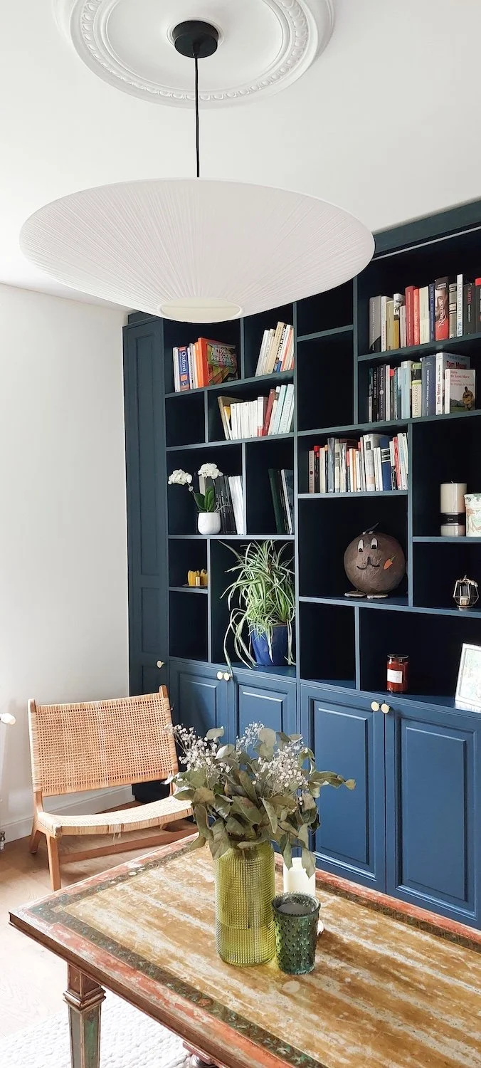 Un salon avec une grande bibliothèque noire, un fauteuil en bois avec un siège en tressage, une table en bois avec des vases de fleurs, et un plafonnier blanc suspendu.