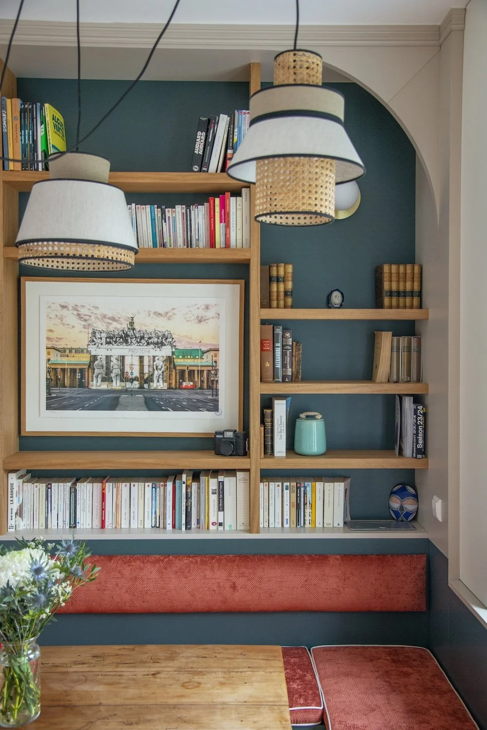 Une bibliothèque en bois avec des étagères remplies de livres, une photo encadrée représentant un monument, deux lampes suspendues au plafond, un vase turquoise, une camera noire, des objets décoratifs, un bouquet de fleurs dans un vase en verre, une nappe rouge en tissu sur une table en bois.