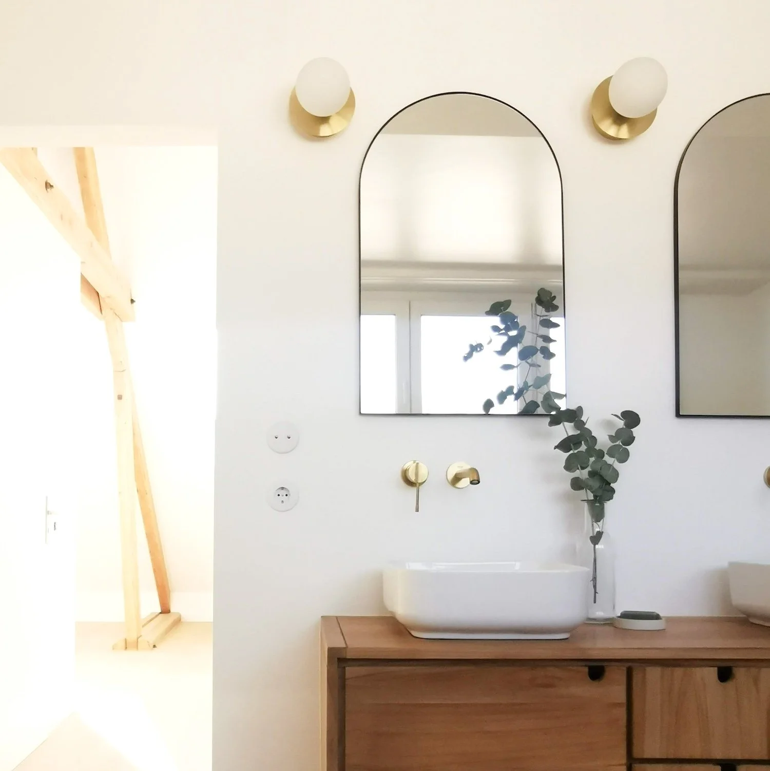 Salle de bains minimaliste avec miroir arqué, lavabo blanc, meubles en bois, plante verte dans un vase, lumières murales blanches