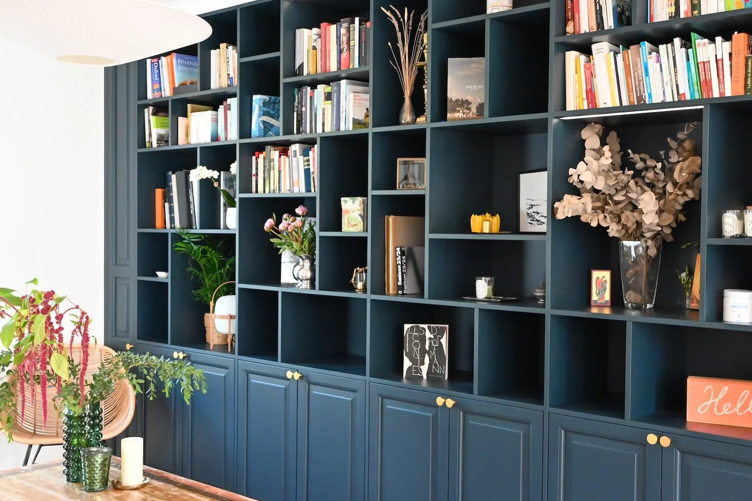 Bibliothèque bleue avec des étagères remplies de livres, décorations florales et objets divers dans une pièce bien éclairée.