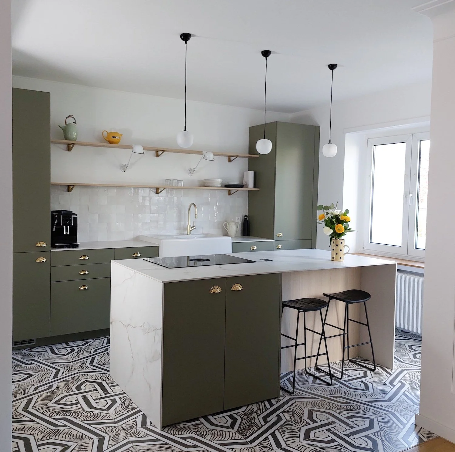 Cuisine moderne avec un îlot central en marbre blanc, armoires vertes, étagères en bois, luminaires suspendus, fenêtre, et planche à découper avec un vase de fleurs.
