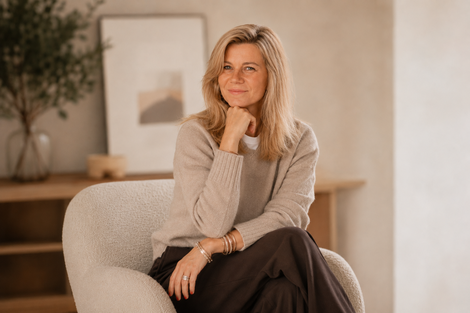 Portrait de Dorothée Vincey assise dans un fauteuil, souriante, portant un Pull beige et des bracelets en or, dans un intérieur moderne avec une décoration minimaliste
