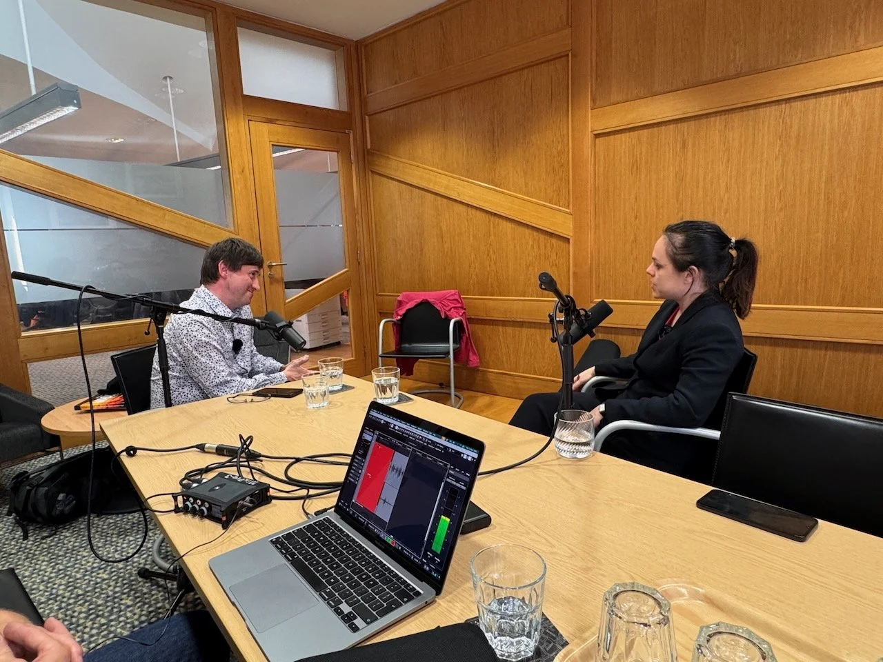 An interview or podcast recording session with two people seated across from each other at a wooden table with microphones. The woman on the right has dark hair tied back, and the man on the left has short brown hair. There are glasses of water and recording equipment on the table.