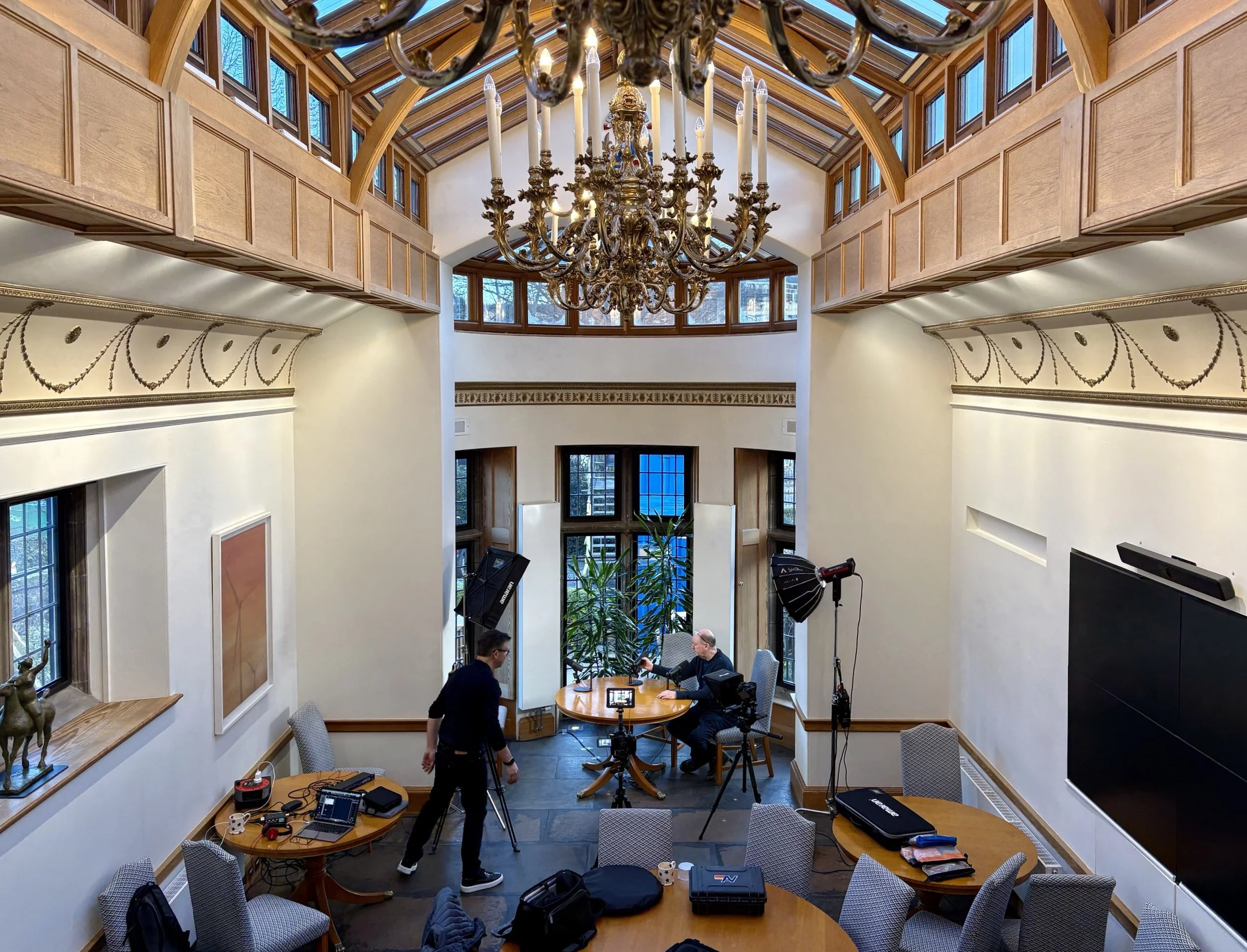 Interior of a room with high wooden ceiling, chandelier, and multiple windows, set up for a video or photo shoot with cameras, lighting equipment, and people preparing.