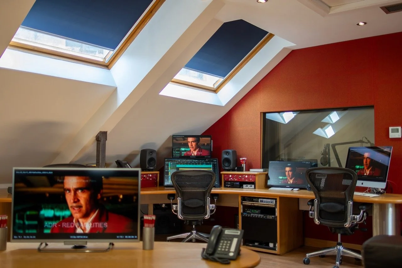 A recording studio with a sloped ceiling featuring skylight windows. The studio has multiple monitors, computers, and audio equipment, with two office chairs facing the workstations. There is a red accent wall, and a large window reflecting the studio equipment.