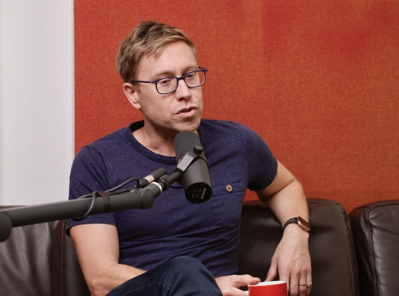 A man with glasses, wearing a navy blue shirt, sitting on a brown leather couch, speaking into a microphone during a podcast or interview, with a red and white background.