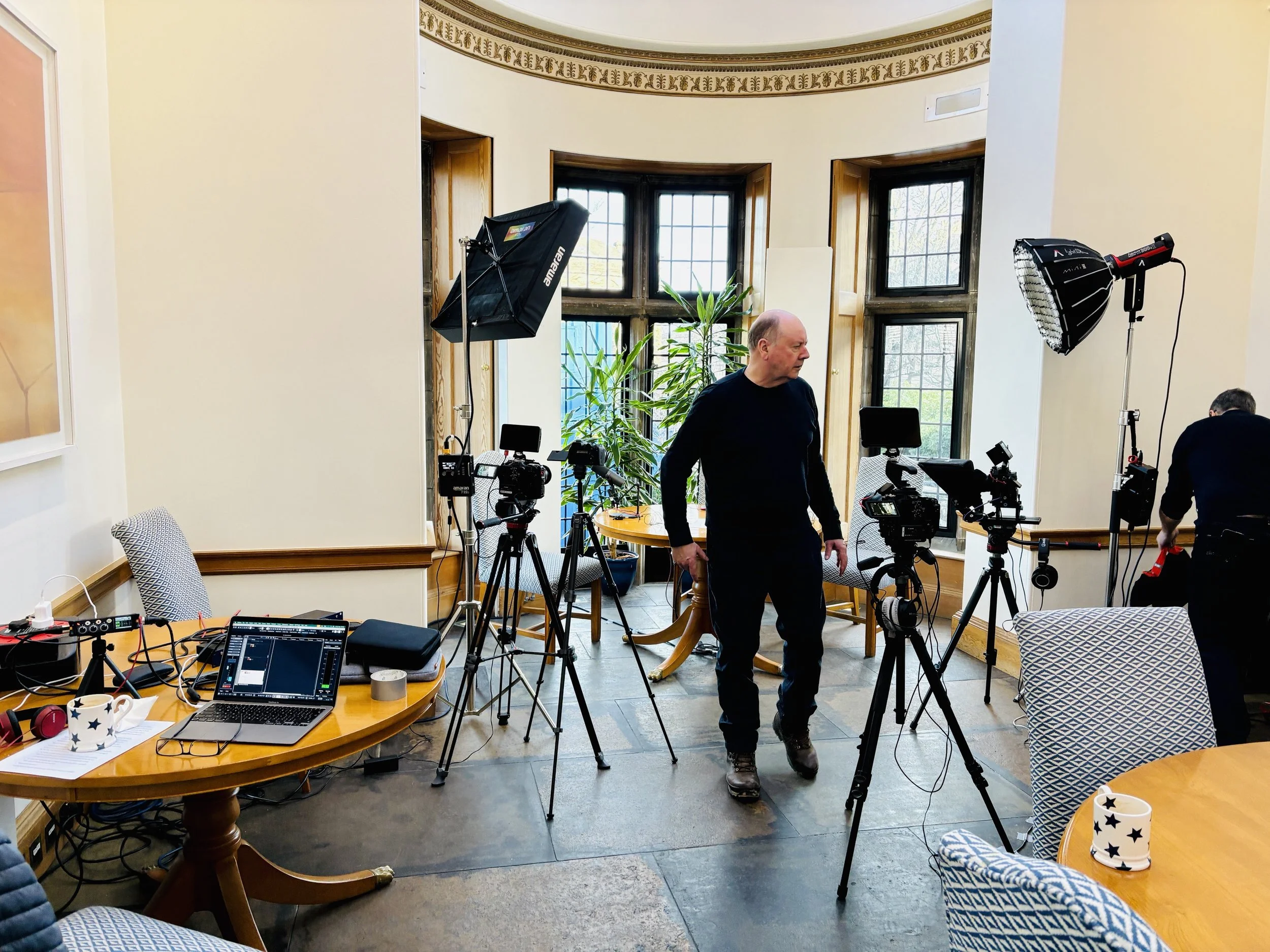 A filming setup in a bright room with multiple cameras and lighting equipment, someone preparing the equipment, and a table with a laptop and coffee mugs.