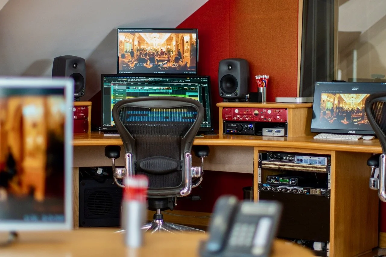 Inside a recording studio control room with a computer monitor displaying audio editing software, a desktop computer, and various audio equipment, including speakers, microphones, and editing tools.