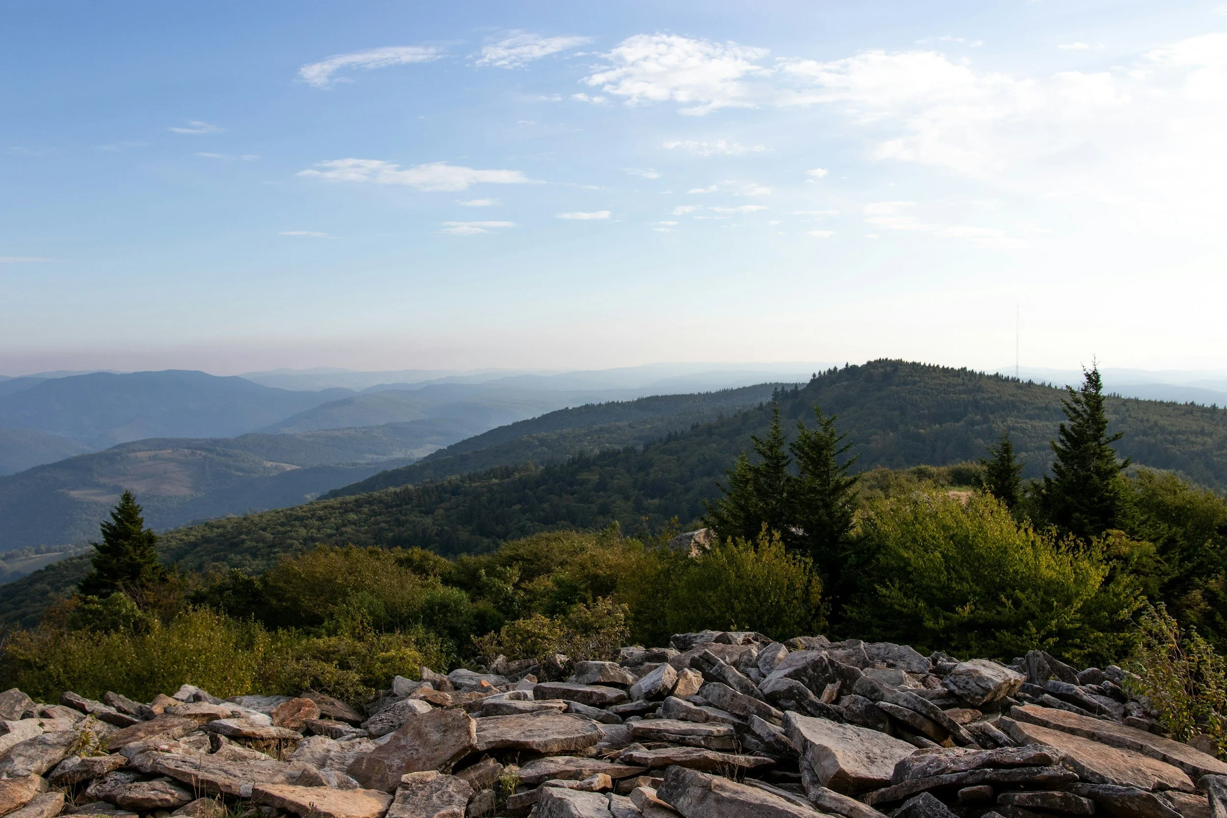 Mountain landscape with rocky foreground, green forested hills, and a clear sky with some scattered clouds.