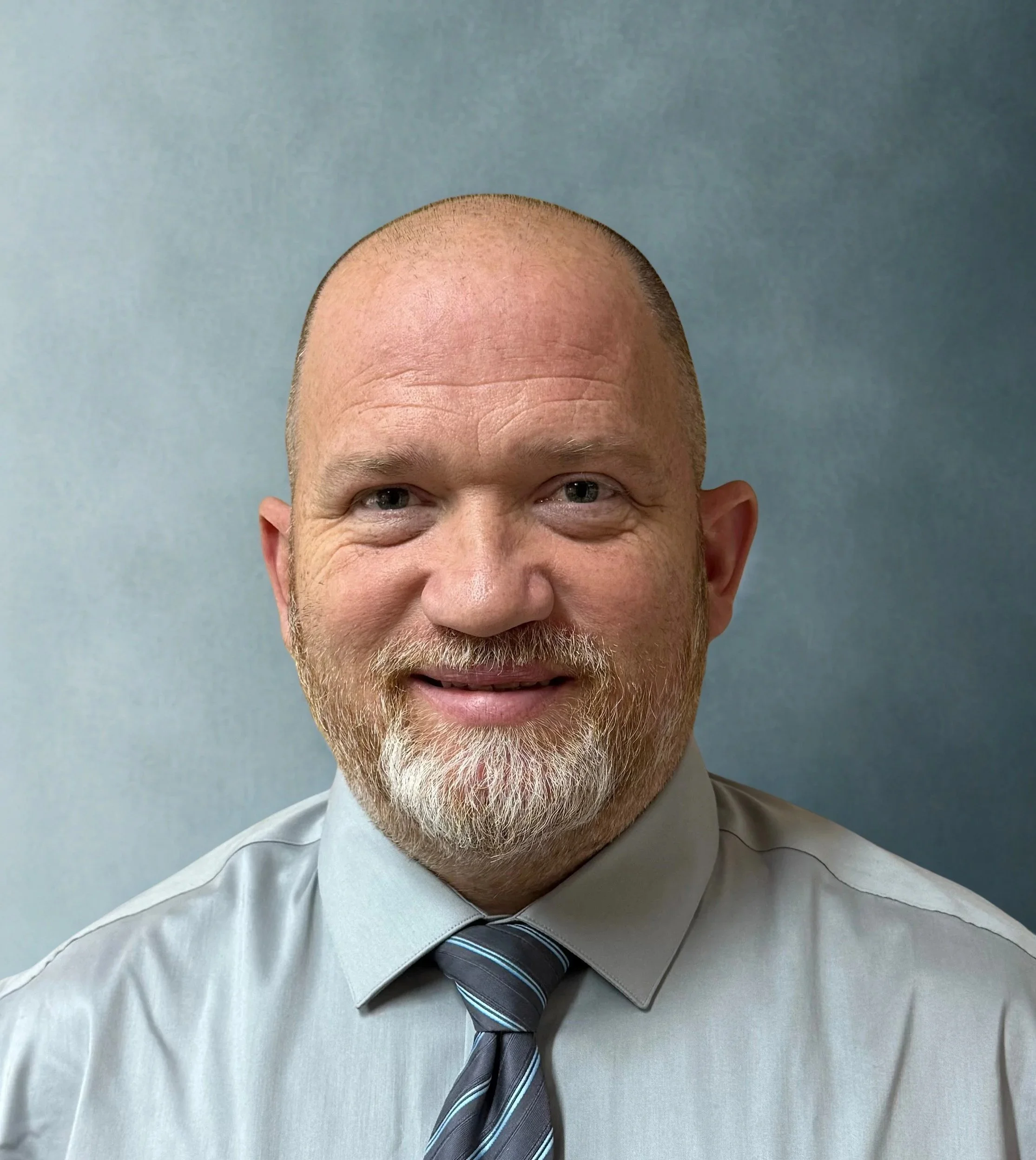 A man with a shaved head and beard wearing a gray dress shirt and a striped tie, smiling against a plain gray background.