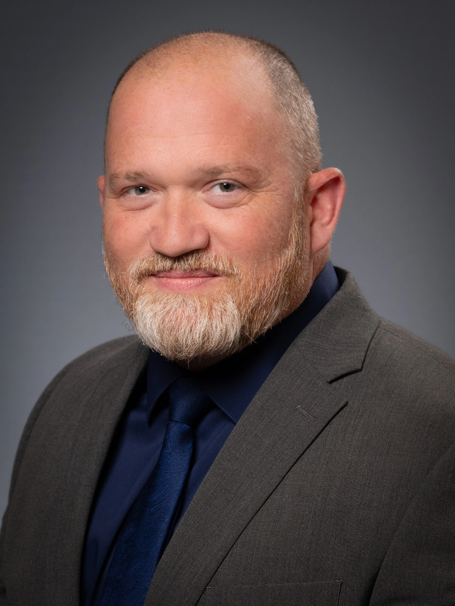 A headshot of a man with a bald head, gray beard, and light skin, wearing a dark suit, dark shirt, and blue tie, against a gray background.