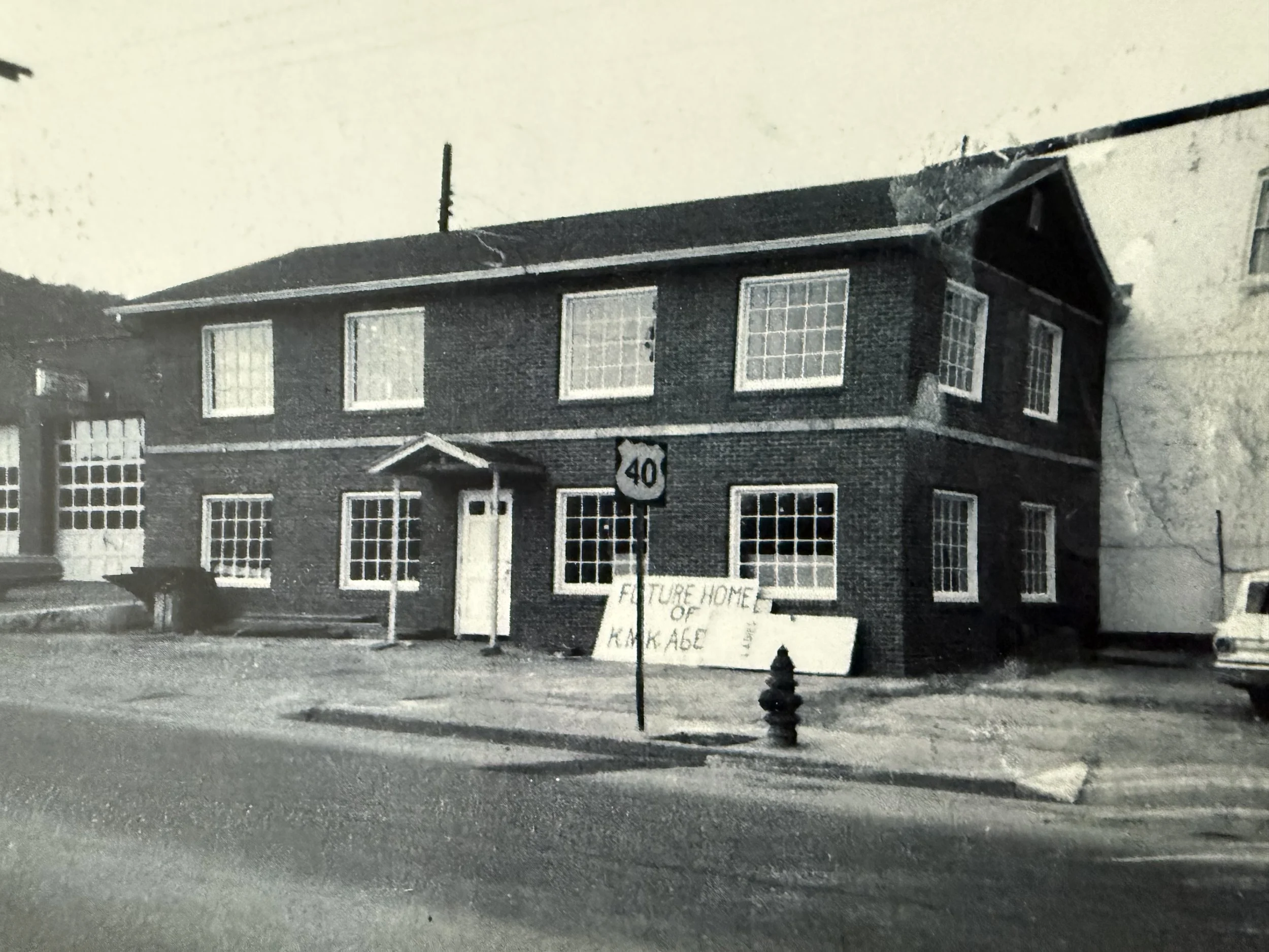 Black and white photograph of a two-story house with multiple windows, a small porch with a porch roof, and a house number 40 on a sign in front. There is a sign on the lawn that reads 'Future home of Karam'. A street sign indicating the 40 block is in the foreground.