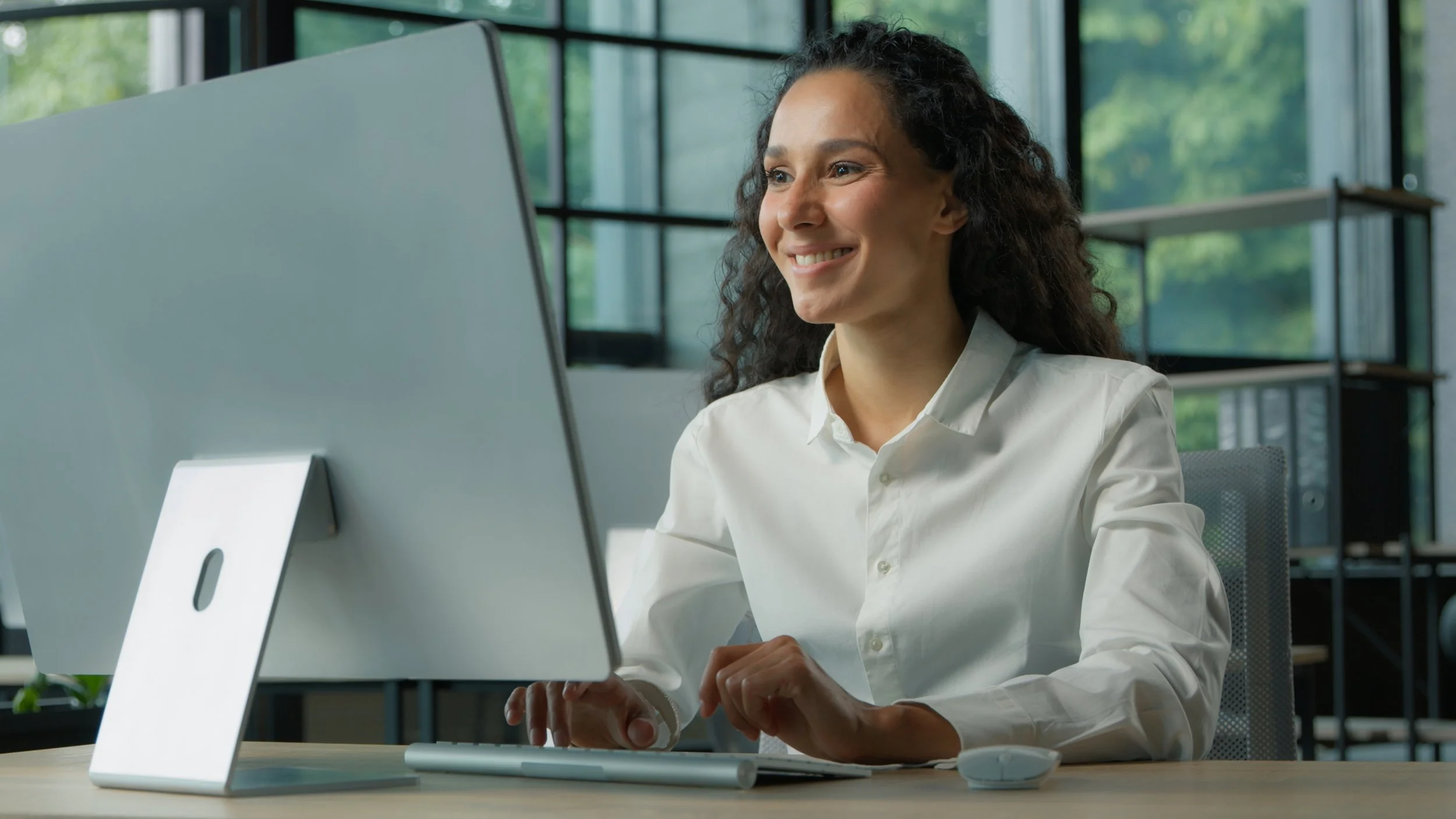 A woman working at a computer in an office with large windows showing trees outside.