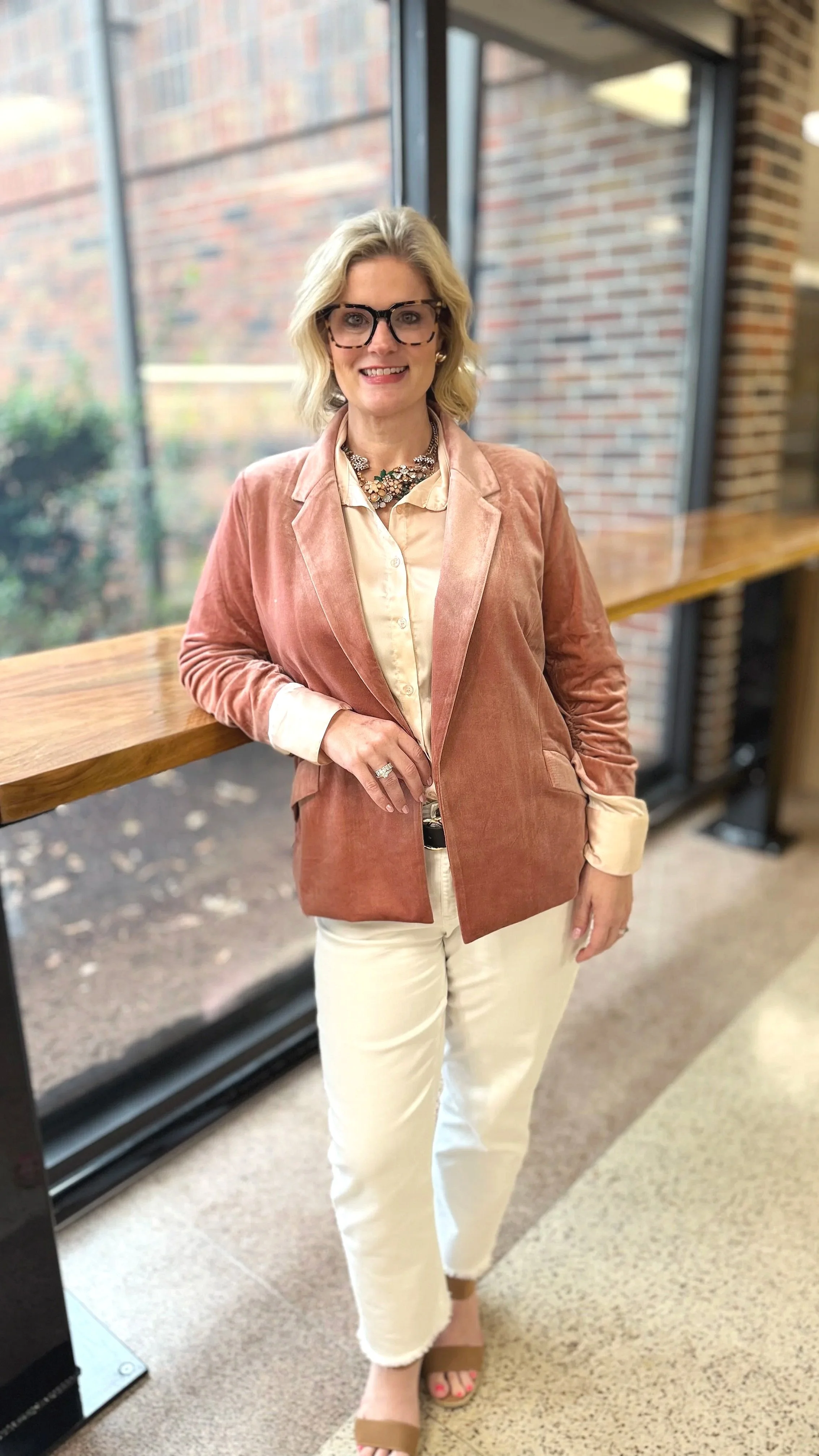 Woman with blonde hair and glasses standing indoors near a large window with brick wall outside. She is wearing a peach-colored velvet blazer, a cream-colored blouse, cream pants, tan open-toe shoes, and accessorized with a necklace and rings.