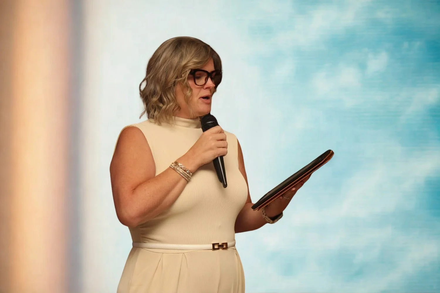 A woman with shoulder-length blonde hair, wearing glasses and a sleeveless beige dress, holding a microphone in one hand and reading from a tablet in the other, standing against a blue and white backdrop.