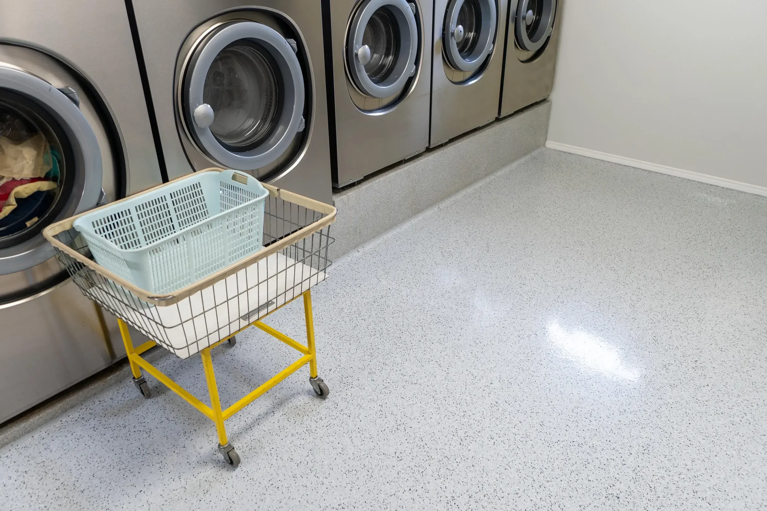 A row of silver commercial washing machines in a laundry room, with a yellow laundry cart with white and beige baskets in front.