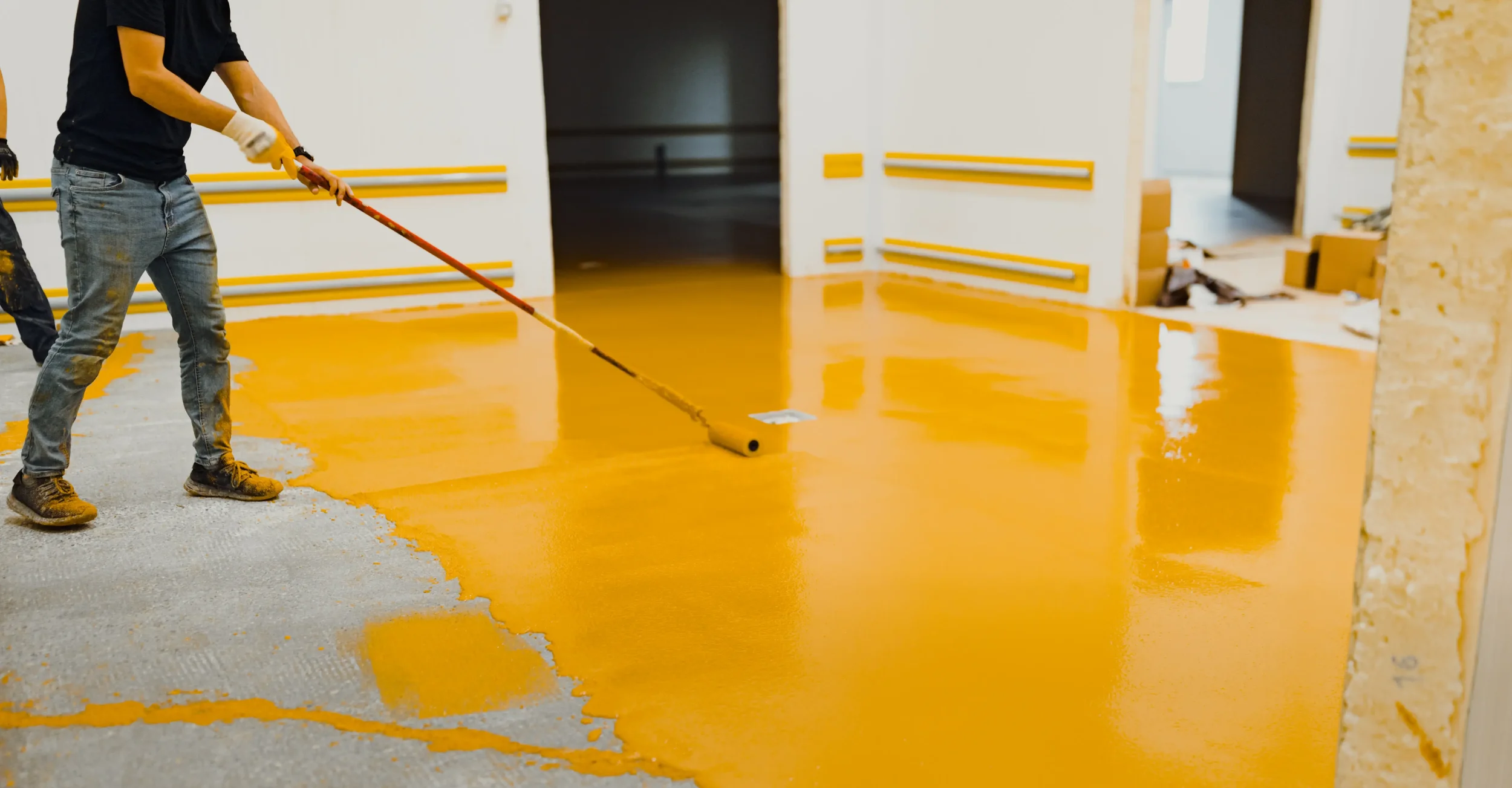 Person rolling yellow epoxy paint on a concrete floor in an indoor construction site.