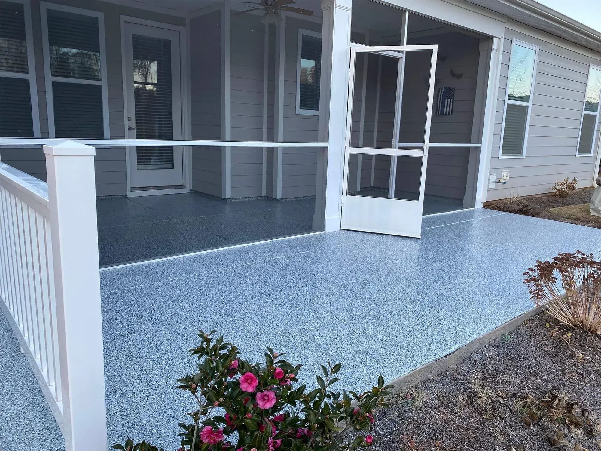A screened porch with a door, windows, and a white railing in front of a house with gray siding, small bushes, and blooming pink flowers.