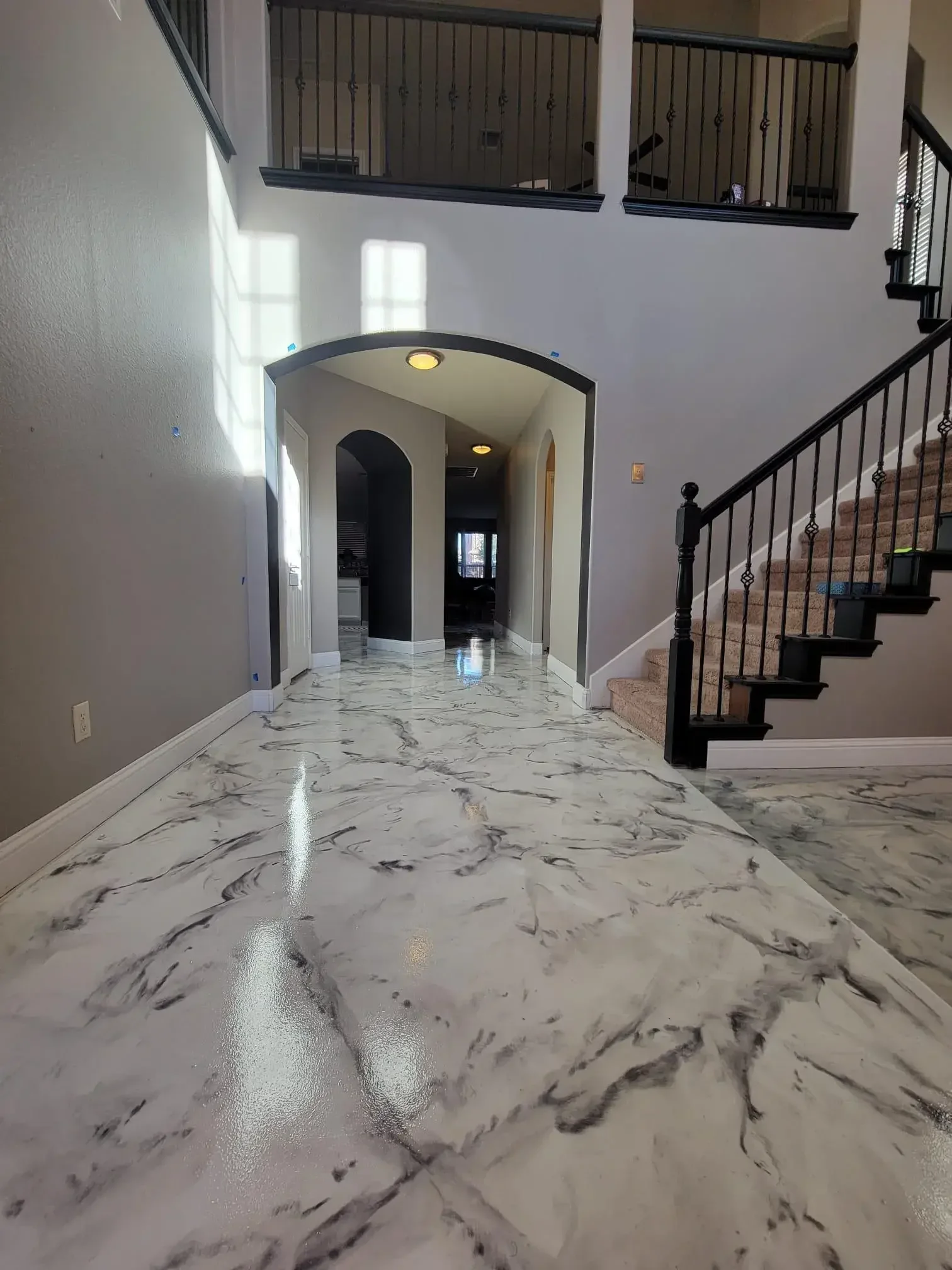Interior view of a home showing a hallway with white marble flooring with black and gray veining, light gray walls, an arched doorway, a staircase with beige carpet and black metal railing, and upstairs balcony with black metal railing. Sunlight cast