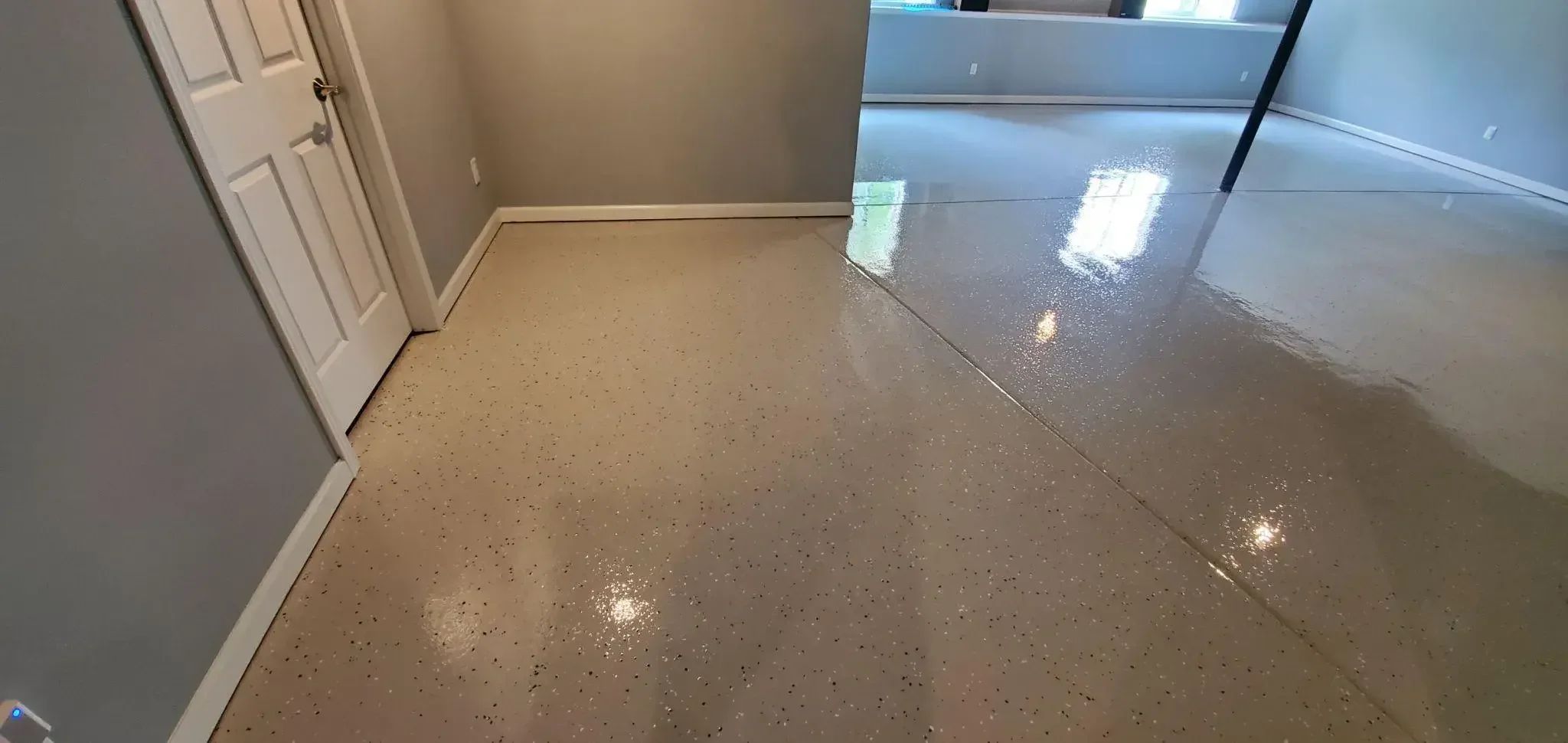 Interior view of a room with a polished, light-colored concrete floor, a closed white door on the left, and a large window in the background. The floor transitions from a matte to a shiny finish.