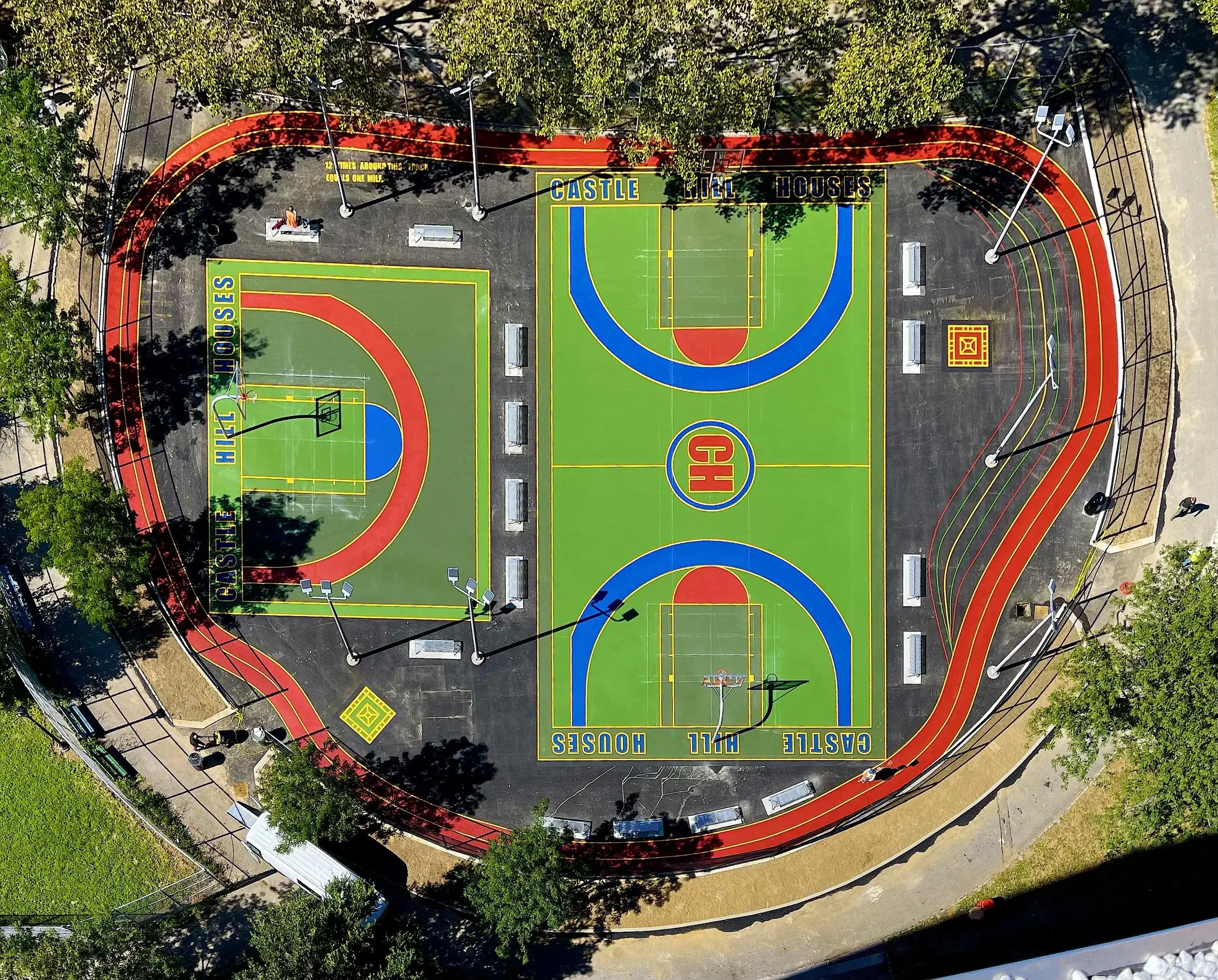 An aerial view of a colorful basketball court with bright green, blue, red, and yellow markings, surrounded by a red running track, and with some trees and benches around it.