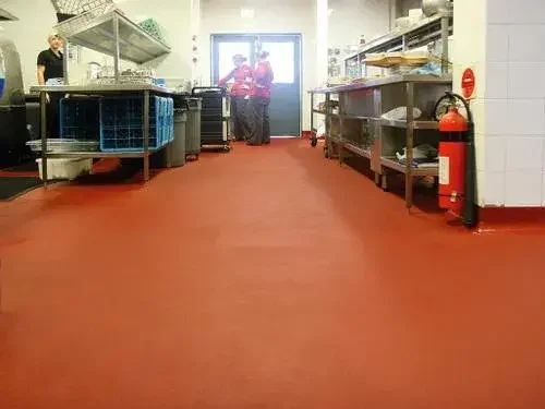 Commercial kitchen with metal shelving, food prep areas, and fire extinguisher, viewed from red flooring towards back door with people inside.