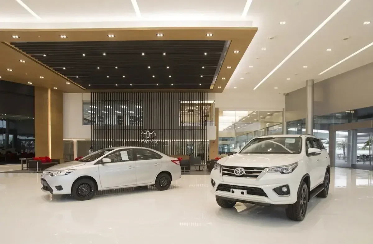 Inside a car dealership showroom with two white cars on display, a sedan on the left and an SUV on the right, with modern interior design and large windows.