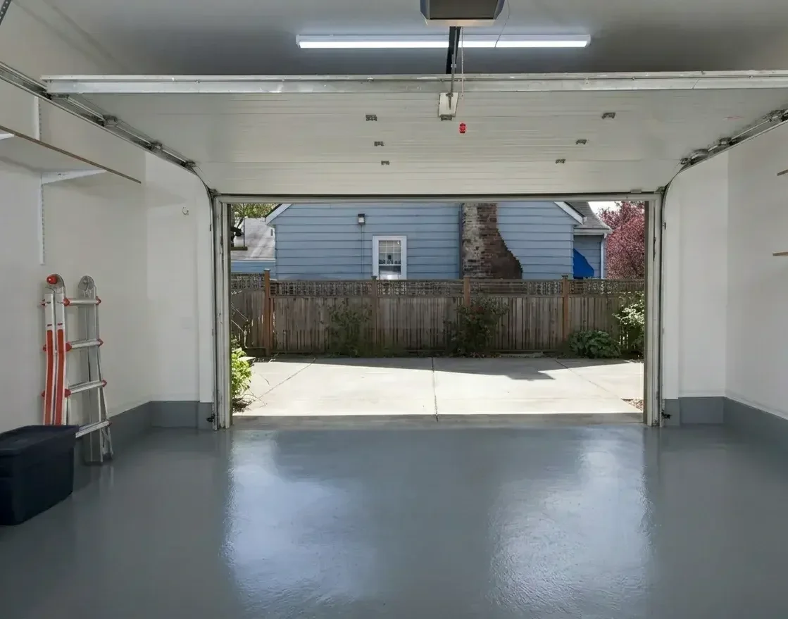 Inside a clean, empty garage with an open door showing a backyard with a fence, house, and driveway.