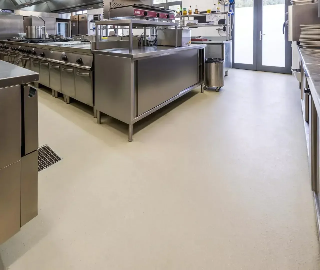 Empty commercial kitchen with stainless steel appliances and counters, and double glass doors at the back.